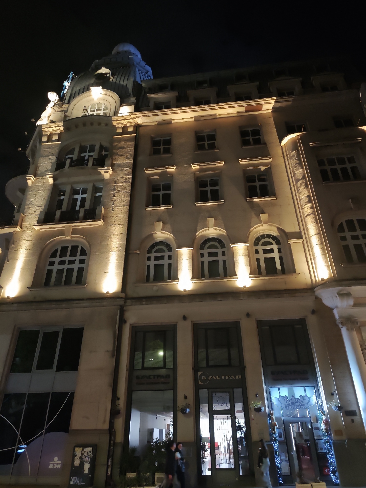This is a night shot of a building in Sofia, Bulgaria. The building is brightly lit with many windows, some arched, some rectangular. On the top of the building, there is a dome-like structure. At the base of the building, there are shopfronts with the Cyrillic word "БУАСТРАД" (BUASTRAD) above two of them. People are visible near the entrance to one of the shops. The overall impression is that of a European city at night.
