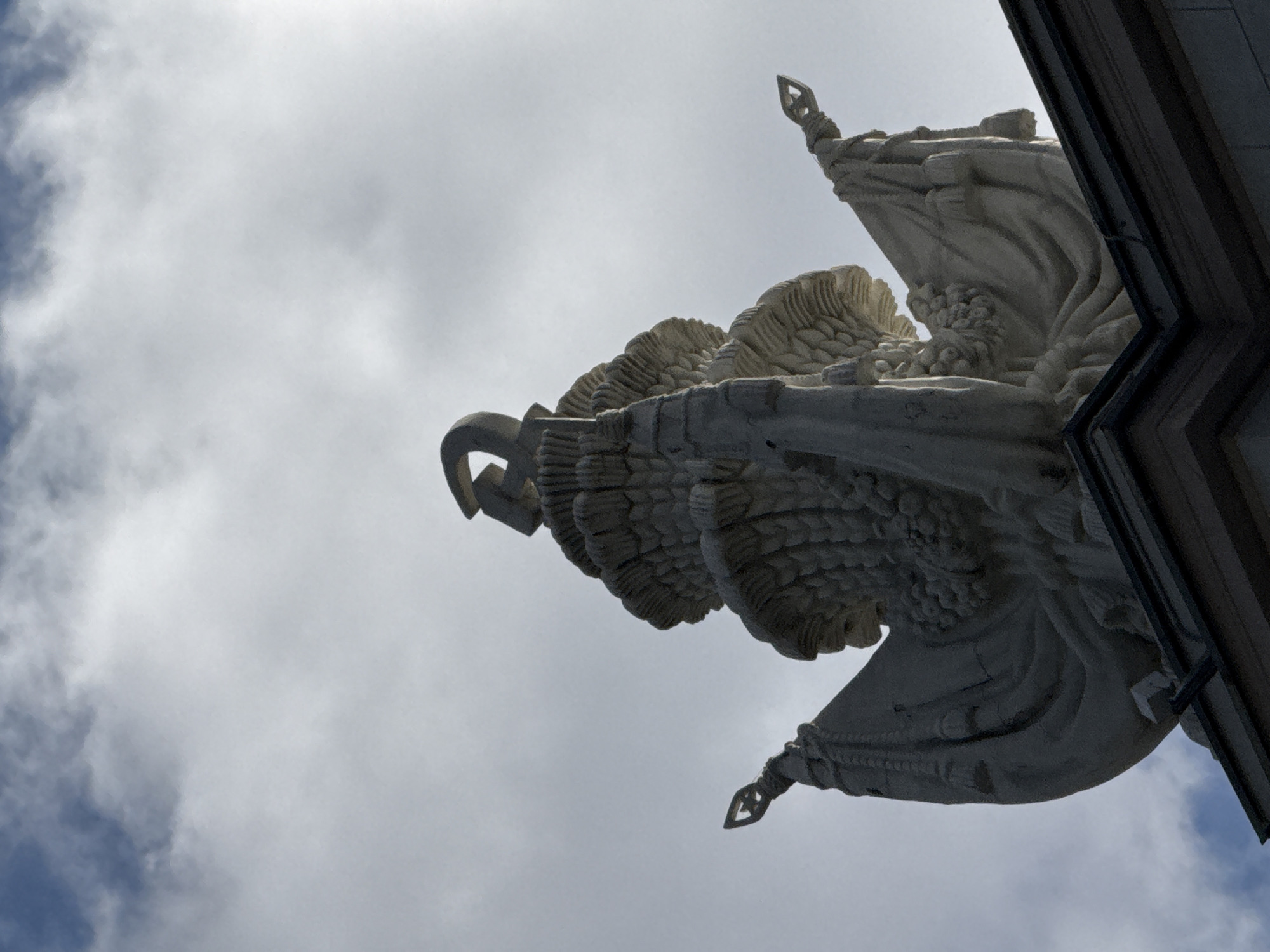 The image shows a close-up, low-angle view of the top of a building in Moscow, Russia, featuring an ornate stone sculpture against a cloudy sky. The sculpture is primarily light grey and appears to be made of stone. It is composed of several layers, resembling a tiered headdress or a stylized bouquet. At the very top, a crescent-shaped object is visible, seemingly part of a finial. Flanking the central sculpture are two draped flags, also rendered in stone, which extend outwards and downwards. The flags are adorned with tassels and have decorative, shield-like finials at their upper points. The sculpture is situated on a dark-colored building, only the very top edge of which is visible in the lower part of the frame. The sky is overcast with patches of bright light suggesting the sun is behind the clouds, creating a dramatic contrast with the sculpture. The overall impression is one of grandeur and historical significance, likely a detail from a prominent architectural landmark in Moscow.