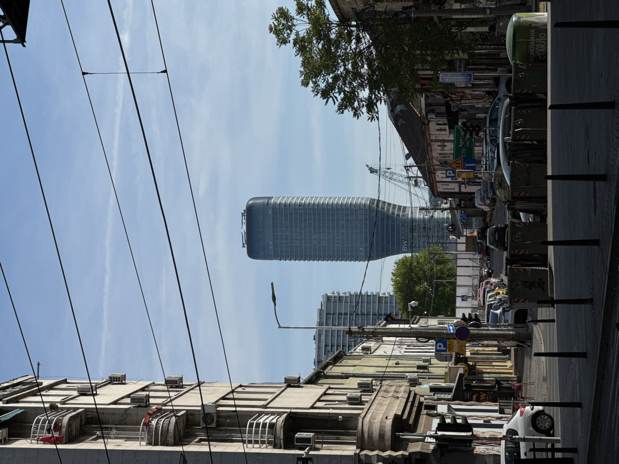 The image captures a street view in Belgrade, Serbia, characterized by an eclectic mix of old and modern architecture. In the foreground, the aged facades of buildings line the street, revealing signs of wear and aging, with air conditioning units and vintage architectural details. Above, a web of power lines cuts across the frame. In the background stands a modern glass skyscraper, the BW Tower, its distinctive shape contrasting with the older buildings. Beyond this tower, another high-rise building is visible. The street below is filled with parked cars, street signs, and waste bins, indicating a busy urban environment. The sky is clear with light clouds, suggesting a sunny day. There are no people immediately visible in the image.