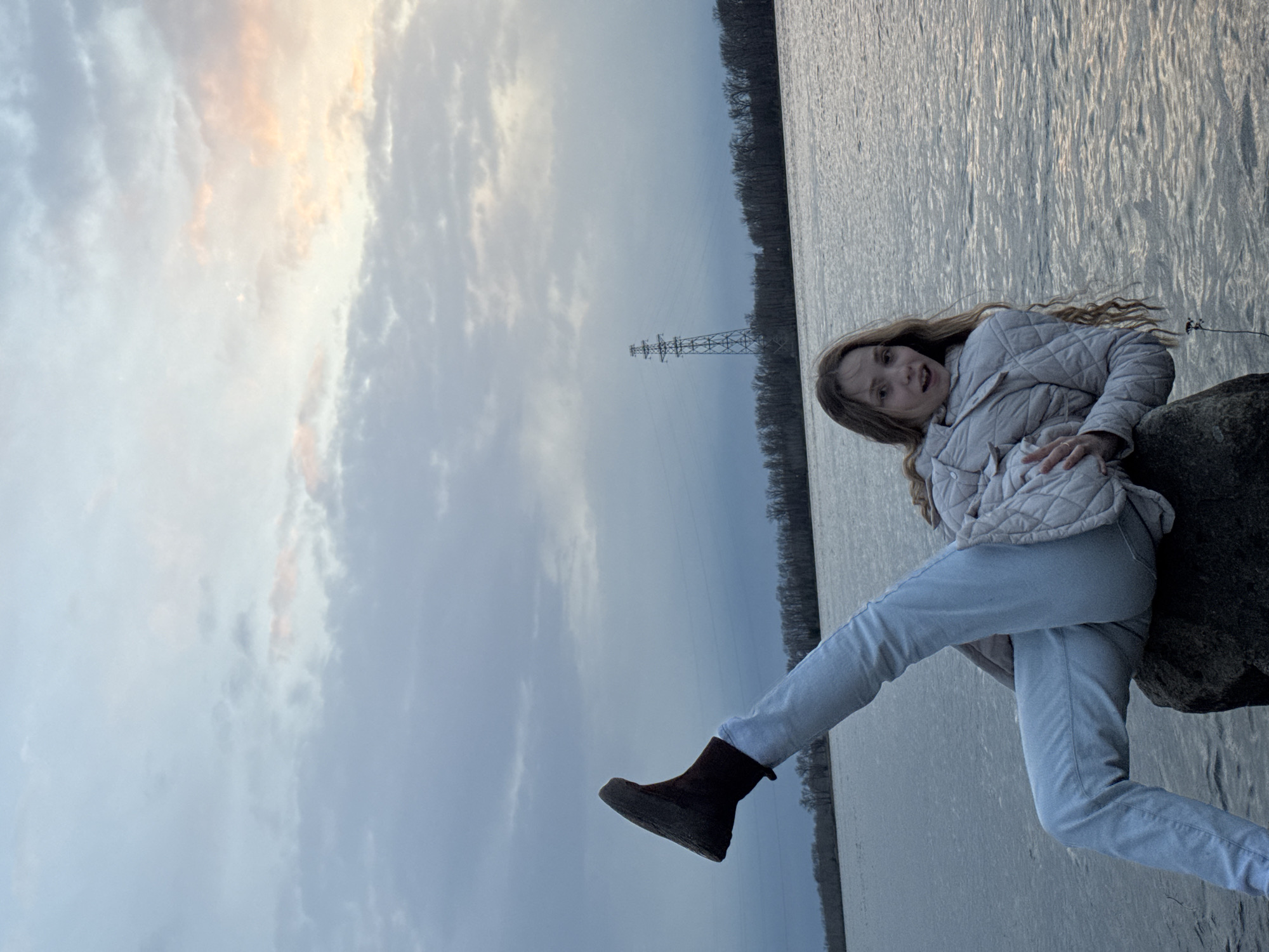 A young girl with long, wavy blonde hair is captured posing playfully outdoors, likely in Chaikovsky, Russia, near a large body of water. She is wearing a light, quilted jacket, light blue pants, and dark boots. Her back leans against a dark rock or boulder located at the edge of the water, and her left leg is lifted in a dynamic, energetic stance. Her mouth is open in an expressive smile or shout, conveying a sense of joy and spontaneity.
The setting features a wide body of water, possibly a lake or river, stretching across the middle and right side of the frame, exhibiting small ripples on its surface. Beyond the water, a dense treeline forms a dark horizontal band on the distant bank. Above the treeline, a prominent metal electricity pylon or transmission tower stands silhouetted against the sky. The sky itself, particularly in the upper left portion of the image, displays a mix of soft, cool grey clouds and warmer, faint orange and pink hues, suggesting the scene takes place during either a late afternoon sunset or an early morning sunrise. The lighting is diffused, indicating an overcast or partly cloudy day. No text is visible in the image.