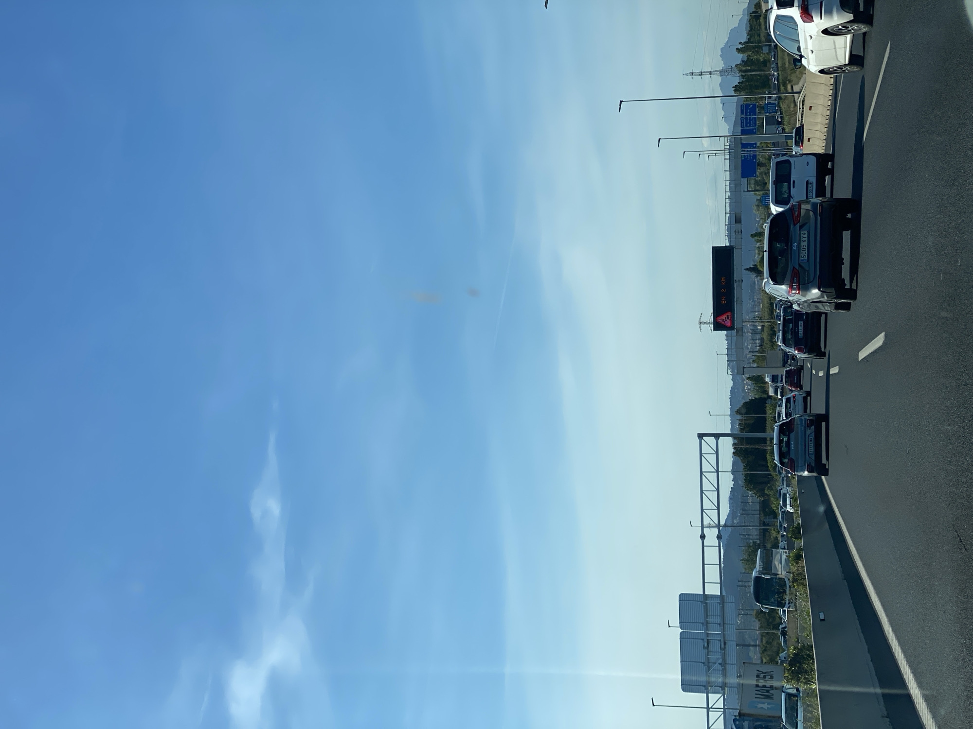 The image depicts a traffic jam on a highway in Sant Joan Despí, Spain. A line of vehicles is visible, including cars, vans, and possibly trucks, all stationary or moving very slowly. The sky is blue with some clouds. Overhead signs and highway lighting are visible alongside the road. Some of the vehicles' license plates are visible. In the background, there's a landscape with hills and possibly some industrial or commercial structures.