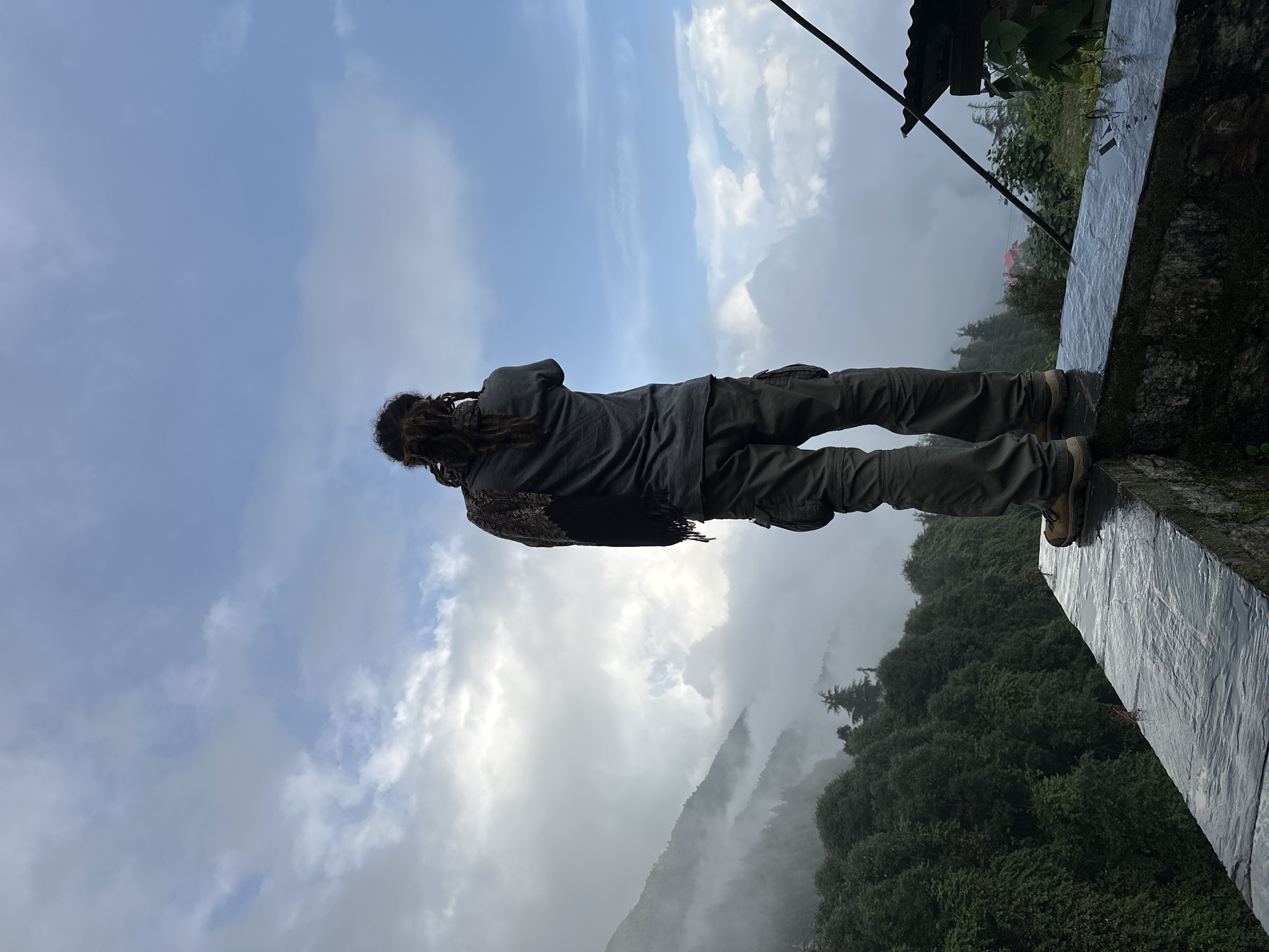 An adult individual with long dreadlocks stands on a stone ledge, facing away from the camera and looking out at a misty mountain landscape. The person is wearing a grey t-shirt, olive-green cargo pants, and brown hiking boots. A patterned scarf is draped around their shoulders. The background features lush green trees, rolling hills partially obscured by thick fog, and a bright blue sky with scattered white clouds. A small red-roofed building is visible in the distance. The stone ledge appears wet, possibly from rain. The scene is set in Haini, India.