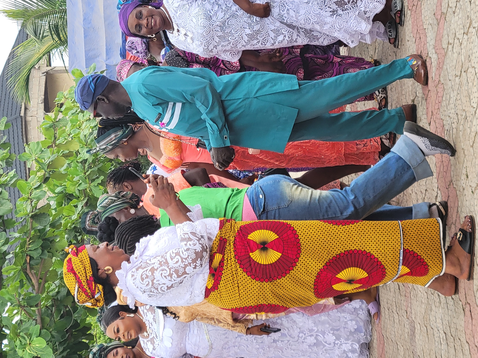The image captures a group of people lined up outdoors. The location is likely Suleja, Nigeria, based on attire and the presence of trees. The subjects are individuals, primarily women, dressed in a variety of traditional African attire, including bright colors, patterns, and headwraps. A man in a teal suit and blue hat stands among them.
Some individuals are interacting with phones. The environment features trees and vegetation, and a paved ground. There is a blue and white striped awning in the background.