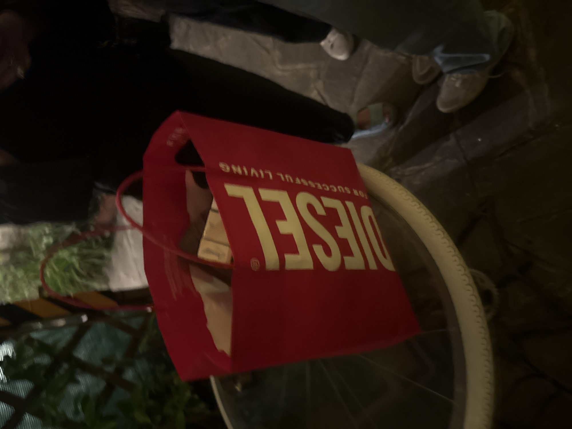 The image shows a red Diesel shopping bag resting on a bicycle wheel. In the background, two people are standing on a stone street. The image suggests the location is in Milan, Italy, based on contextual clues.