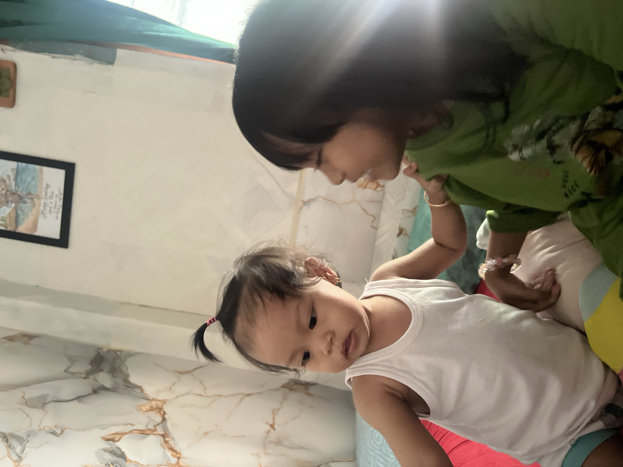The image shows two young girls indoors, likely in a residential setting within Kecamatan Pati, Indonesia. The girl on the left, appearing to be a toddler, wears a white tank top and has her dark hair pulled up in a small ponytail with a pink hair tie. She has a gold hoop earring in her left ear. The girl on the right, who appears older, has dark hair and is wearing a green long-sleeved shirt. Her face is partially obscured by glare from the lighting. The girls are sitting or lying on a bed, indicated by the presence of pillows and blankets in various colors, including red, beige, and light blue. In the background, the wall has a marbled pattern with golden veins. A framed picture hangs on the wall, and a green curtain is visible to the right. The lighting suggests it is daytime, with a strong light source causing significant glare, particularly on the right side of the image. The overall scene depicts a casual, everyday moment between two children in a home environment.