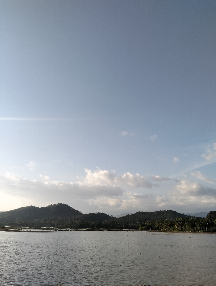 The image depicts a serene landscape. Still water fills the foreground, reflecting the sky above. Behind the water, low, rolling hills covered in lush greenery stretch across the horizon. A clear blue sky dominates the upper portion of the image, punctuated by scattered, soft white clouds. The scene exudes tranquility and peace.