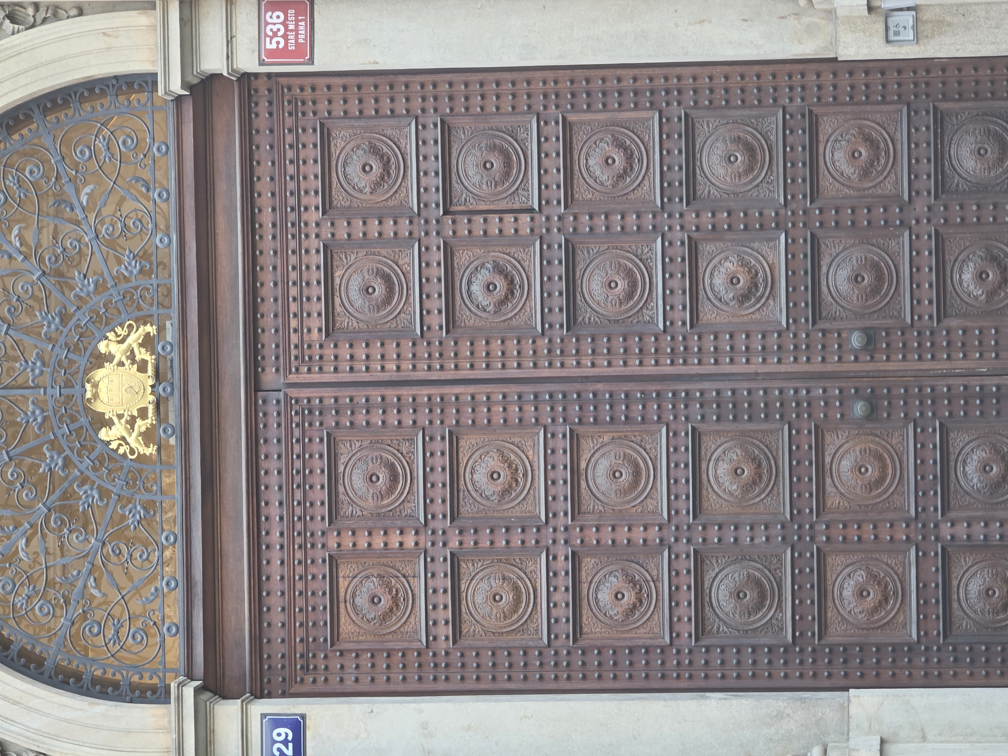 An ornate, dark wooden door stands as the primary subject, adorned with multiple square panels featuring intricate circular, floral-like carvings. Numerous small, rounded studs are embedded into the wood, forming vertical and horizontal rows between and around the panels. The door appears to be split into upper and lower sections, each displaying six carved panels.
To the left of the wooden door is a curved, decorative metal grille or gate with elaborate scrollwork and foliage motifs. Behind this grille, a golden emblem or coat of arms is visible, featuring a lion rampant and a shield. The entire entrance is framed by light-colored stone architectural elements.
Above the wooden door, on the stone facade, a small grey intercom or doorbell unit is mounted.
The scene depicts a static architectural detail, likely an entrance to a building. The lighting suggests daytime.
Visible text provides location cues:
- A red rectangular sign located in the upper right section of the image reads: "536", "STARÉ MĚSTO", and "PRAHA 1", indicating the address is in Prague's Old Town.
- A blue rectangular sign, partially visible in the bottom left, shows the number "29" oriented vertically.