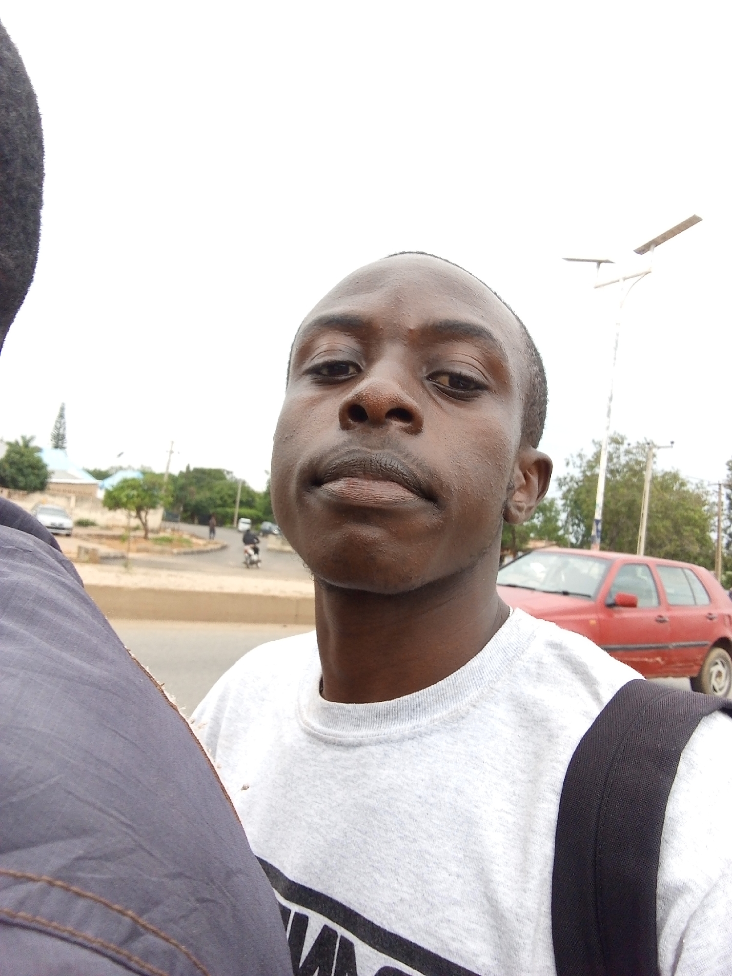 The image depicts a close-up selfie of a young man with dark skin and short black hair, facing the camera. He is wearing a light gray crew-neck t-shirt with a black graphic design on the front and a black backpack strap visible over his right shoulder. The man's expression is neutral, and his head is tilted slightly.
The background suggests an urban environment, likely in Jos, Nigeria. A red hatchback car is parked on the right side of the frame, partially obscured by the man. In the distance, there are trees, buildings, a streetlamp, and what appears to be a dirt road or unpaved area. A few other vehicles and people are faintly visible in the background, indicating a public space. The sky is overcast, contributing to a diffused lighting condition. The overall impression is of a casual, everyday moment captured outdoors.