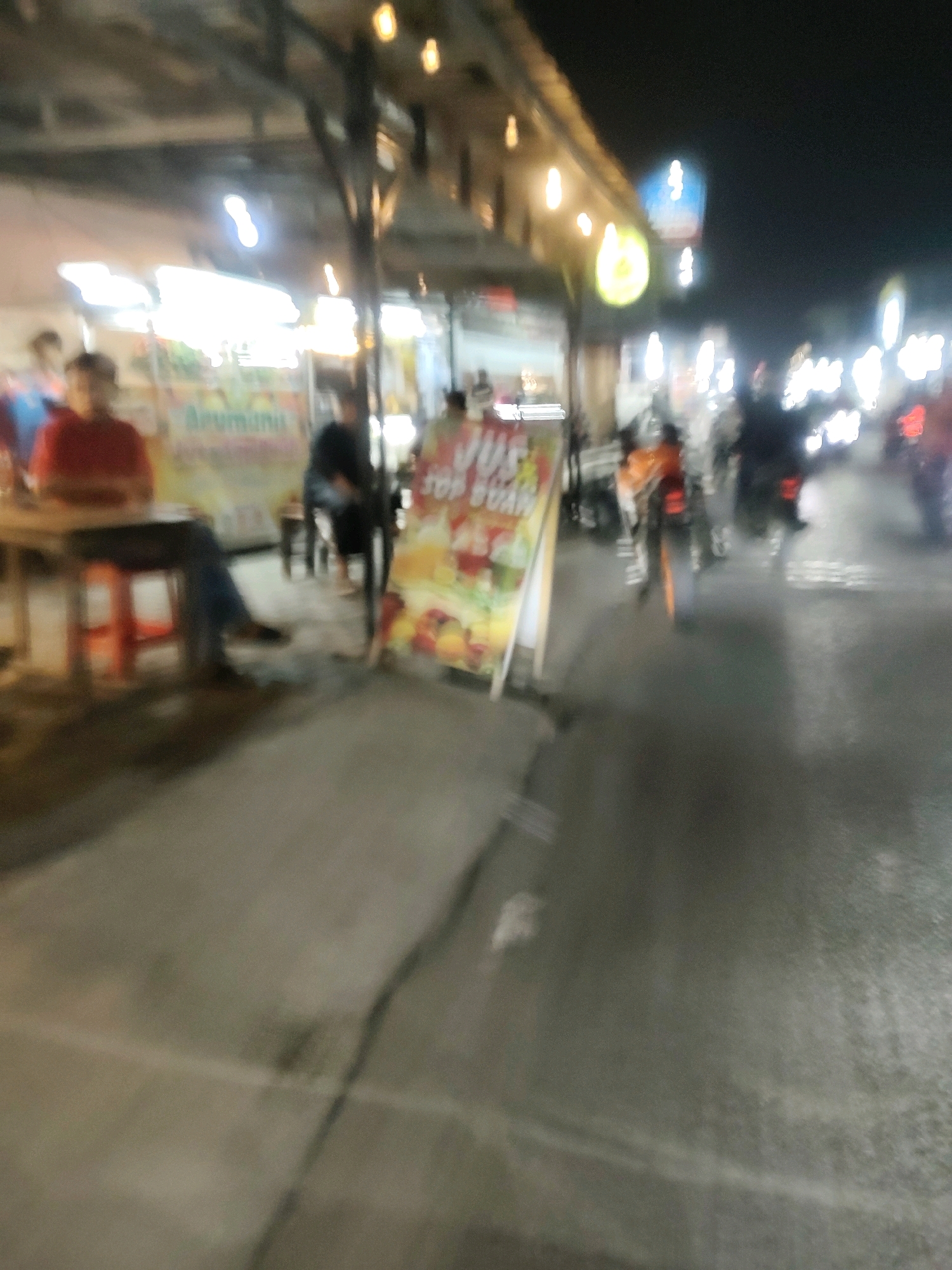 The image depicts an outdoor scene at night, possibly a street food stall or a small market in Mekarmukti, Indonesia. The overall image is blurry, suggesting motion or a low-light, handheld capture. In the foreground, a person in a red shirt is seated at a table with an orange stool. To their right, a banner or sign is visible, displaying text that appears to be in Indonesian, possibly advertising drinks or food with images of fruits. Several other individuals are present in the background, some walking or standing. A few motorcycles are also visible on the street, with their headlights on. The environment includes a paved road, a sidewalk or pedestrian area, and structures that suggest stalls or shops with hanging lights. The lighting appears to be from artificial sources, contributing to the nighttime ambiance.