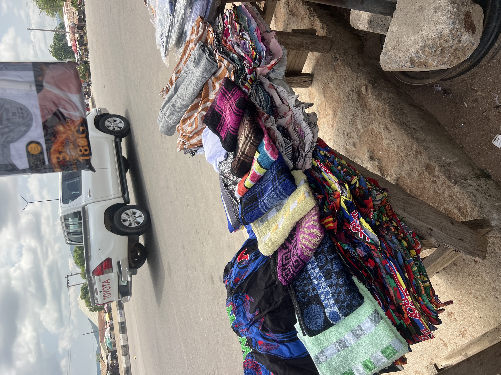 The image depicts an outdoor market scene in Howayi, Nigeria. In the background, a white Toyota pickup truck is parked on the side of a paved road. The truck has "TOYOTA" branding on its tailgate. Beyond the truck, a row of market stalls with various colored awnings are visible, and people can be seen browsing or working at the stalls. The foreground is dominated by a wooden stall laden with a colorful assortment of fabrics and folded textiles, possibly clothing or towels. The stall is set up on a dirt and concrete ground, with some large stones and a black tire rim visible nearby. The sky is cloudy, suggesting an overcast day. The overall impression is one of a busy, functional marketplace.