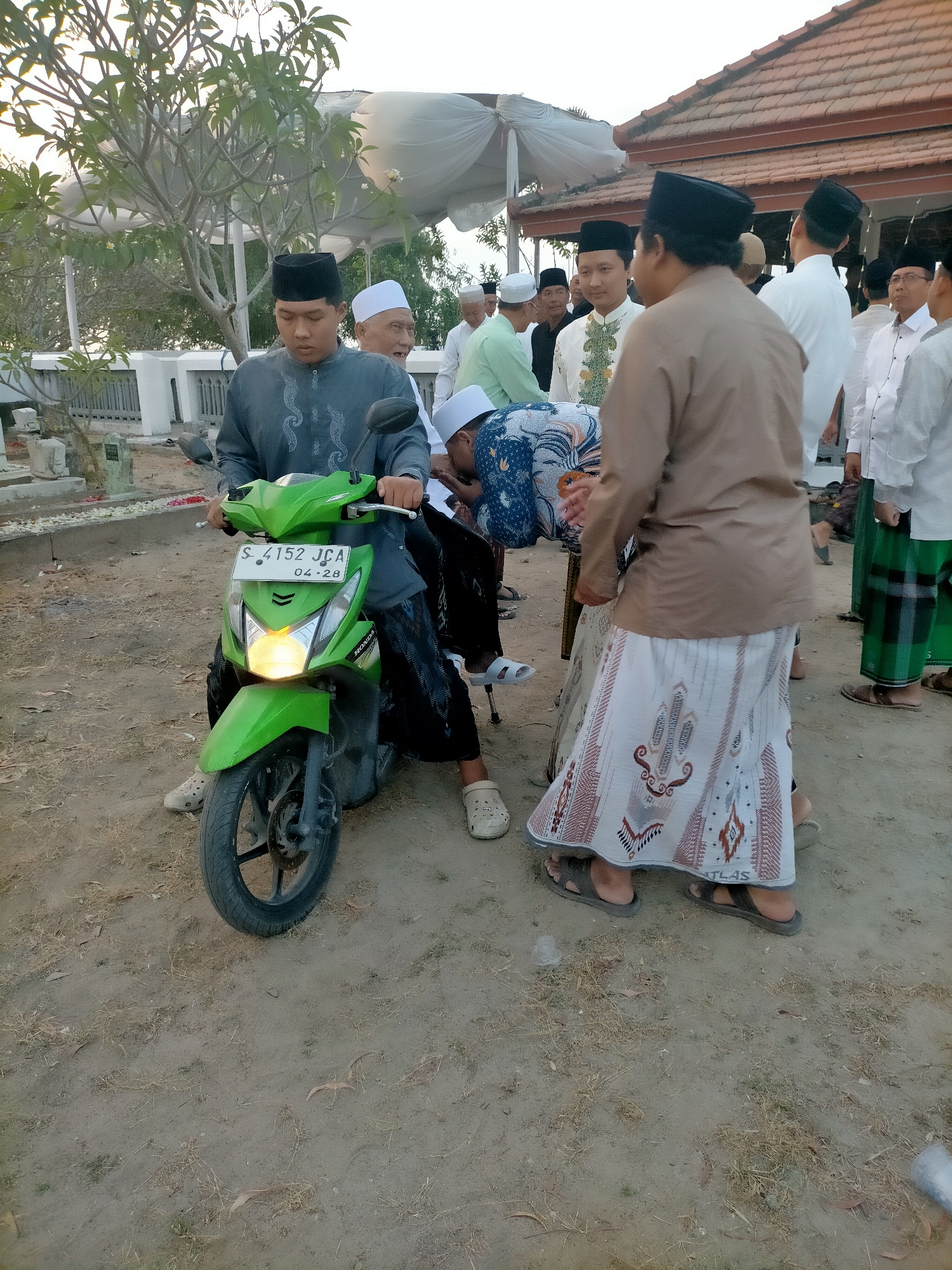 In Kranji, Indonesia, a group of Muslim men, some wearing peci hats, gather outdoors, possibly for a religious event. A young man in a grey long-sleeved shirt and black peci hat is seated on a bright green scooter, his hands on the handlebars. The scooter's headlight is illuminated, and its license plate reads "S-4152 JCA 04-28". Another man, wearing a light brown long-sleeved shirt and a white sarong with a patterned border, stands to the right of the scooter, facing away from the camera. A man in a blue and white patterned sarong is being carried or assisted by another individual. The ground is dirt with sparse dry grass. In the background, a building with a tiled roof and white drapery is visible, along with trees and what appears to be a cemetery with gravestones. The lighting suggests it is either early morning or late afternoon.