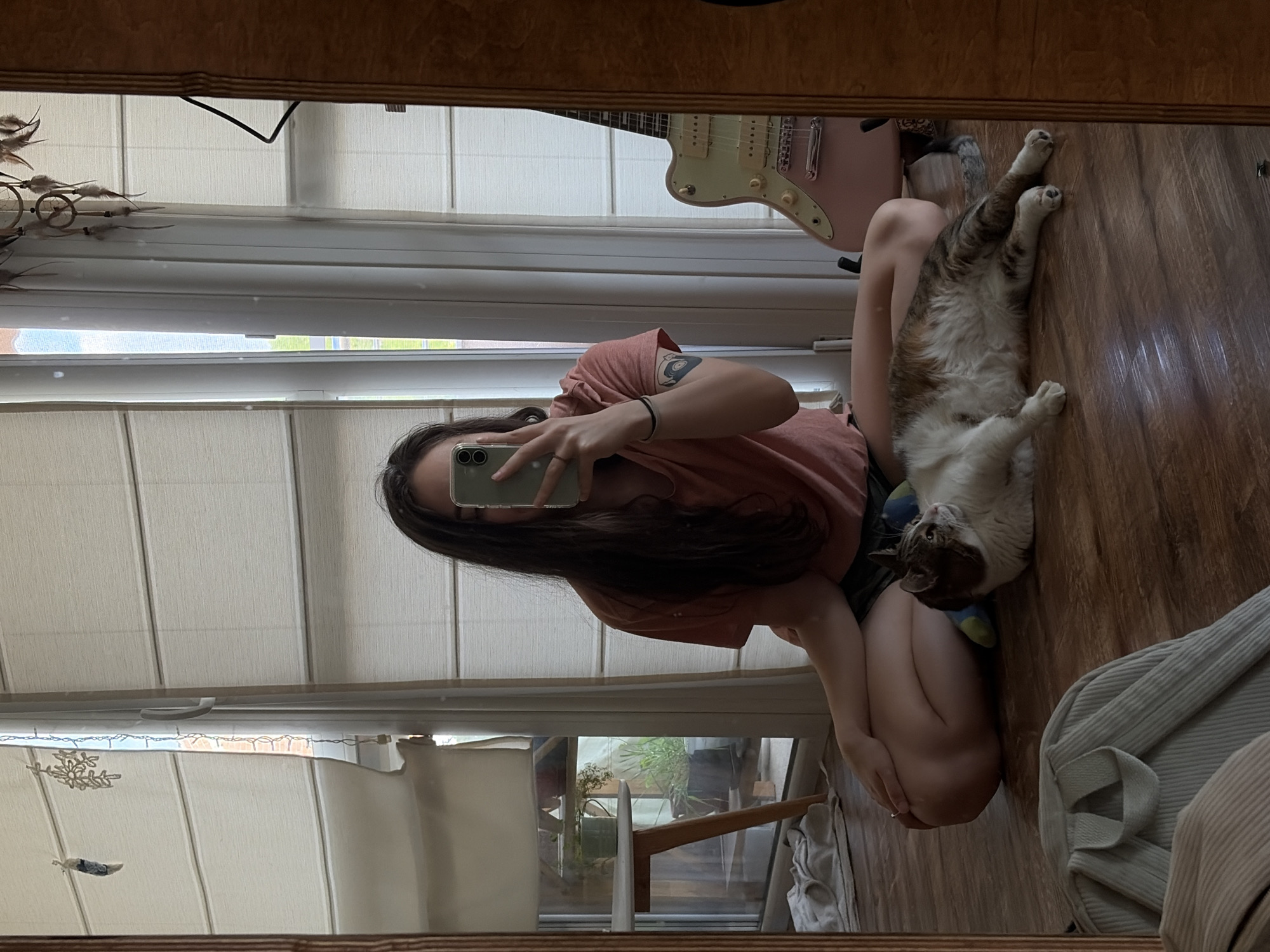 In a room with wooden floors and sheer white blinds covering a large window, a young woman with long, dark hair takes a mirror selfie. She is wearing a pink t-shirt and dark shorts, sitting with her legs crossed and a cat lounging beside her. The cat is a tabby with white markings and appears to be relaxed. Behind the woman, a pink electric guitar is propped up. The reflection in the mirror also shows a small table with plants, and decorative items hanging from the window.