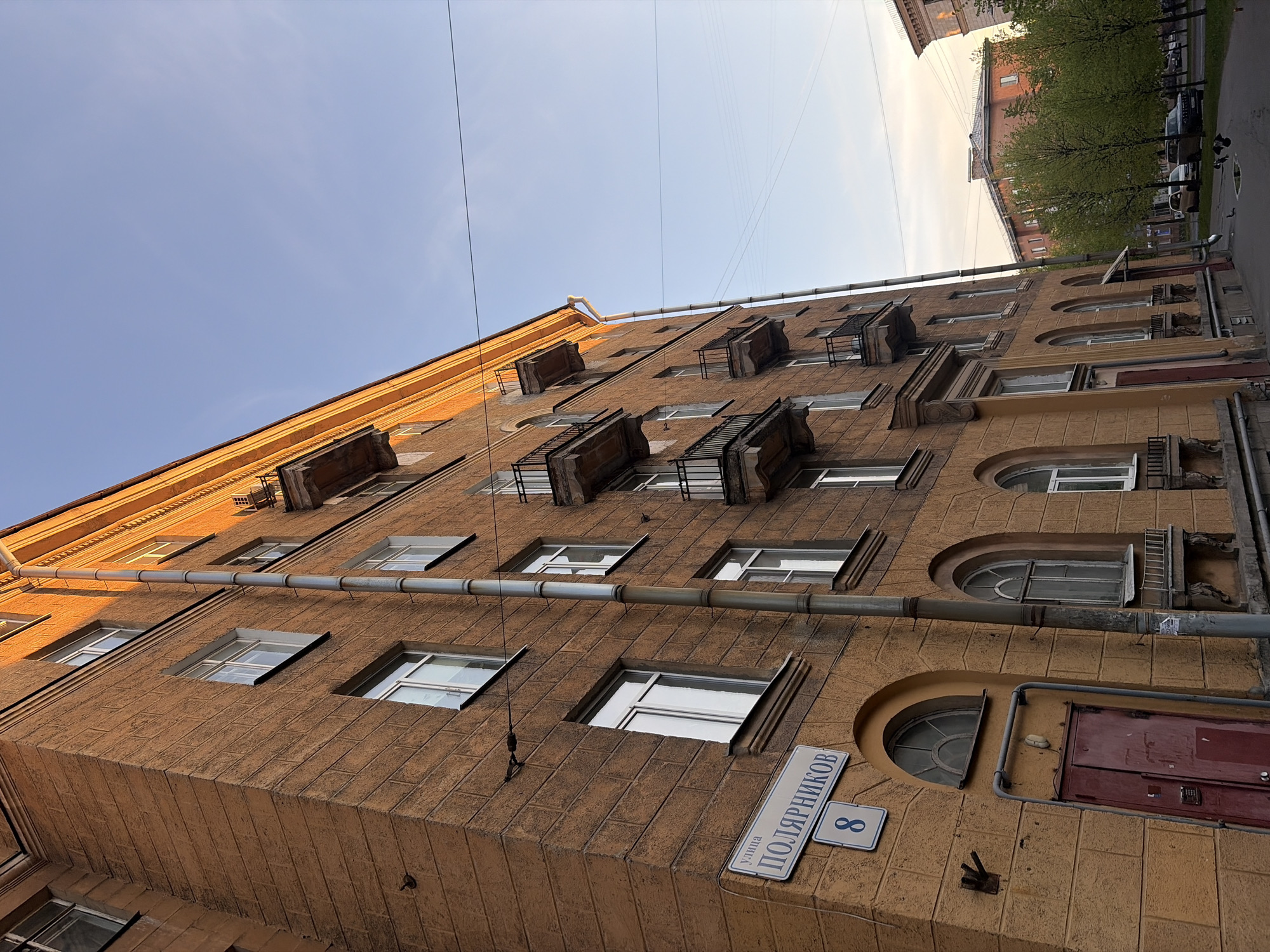 A low-angle shot of a beige building in Saint Petersburg, Russia, reveals rectangular and arched windows, some with balconies. A sign on the building reads "улица ПОЛЯРНИКОВ 8" indicating the street name and building number. Wires run across the top of the building against a blue sky. Part of another building, trees, and parked cars are visible on the right side of the frame.
