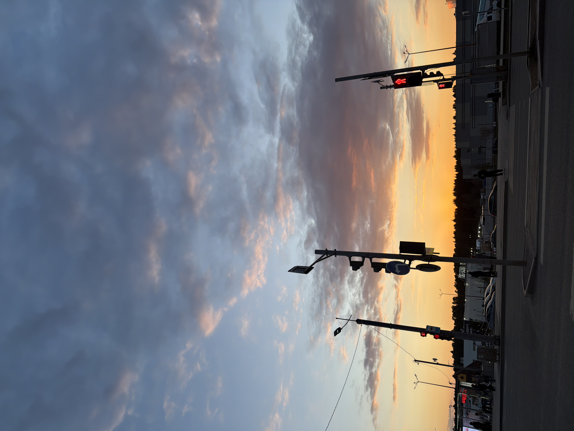 This media file captures an evening street scene at sunset in what appears to be an urban or semi-urban environment. The sky dominates the upper portion of the image, displaying a dramatic blend of dark blue, gray, and warm orange-pink hues from the setting sun, with various clouds scattered across.
In the mid-ground and foreground, several streetlights and traffic light poles are visible. One prominent traffic light shows a red "walking person" signal, indicating that pedestrians should not cross, while another has a solid red circular light. A blue circular road sign with a white arrow pointing right is also attached to a pole. A small rectangular sign on another pole displays the number '5'.
The lower right section of the image shows a paved road with white crosswalk markings. Several vehicles are parked or stopped along the side of the road, appearing mostly as dark silhouettes. A white van is partially visible. A few people are discernible on the pavement, also silhouetted against the brighter background, with one person standing near the crosswalk.
In the background, a line of dark trees forms a stark silhouette against the bright orange horizon, separating the street level from distant buildings. These buildings, also largely silhouetted, show some visible Cyrillic text. One building clearly displays "ПЕРВЫЙ МАГАЗИН" (First Store) and below it "ВЫДАЧА ЗАКАЗА" (Order Collection/Issuance) and "ШИНОМОНТАЖ" (Tire Fitting/Repair). Another sign on a different structure reads "100 МАГАЗИН" (100 Store).
The overall impression is a peaceful evening cityscape, characterized by the beautiful sunset and the usual activities of a street scene.