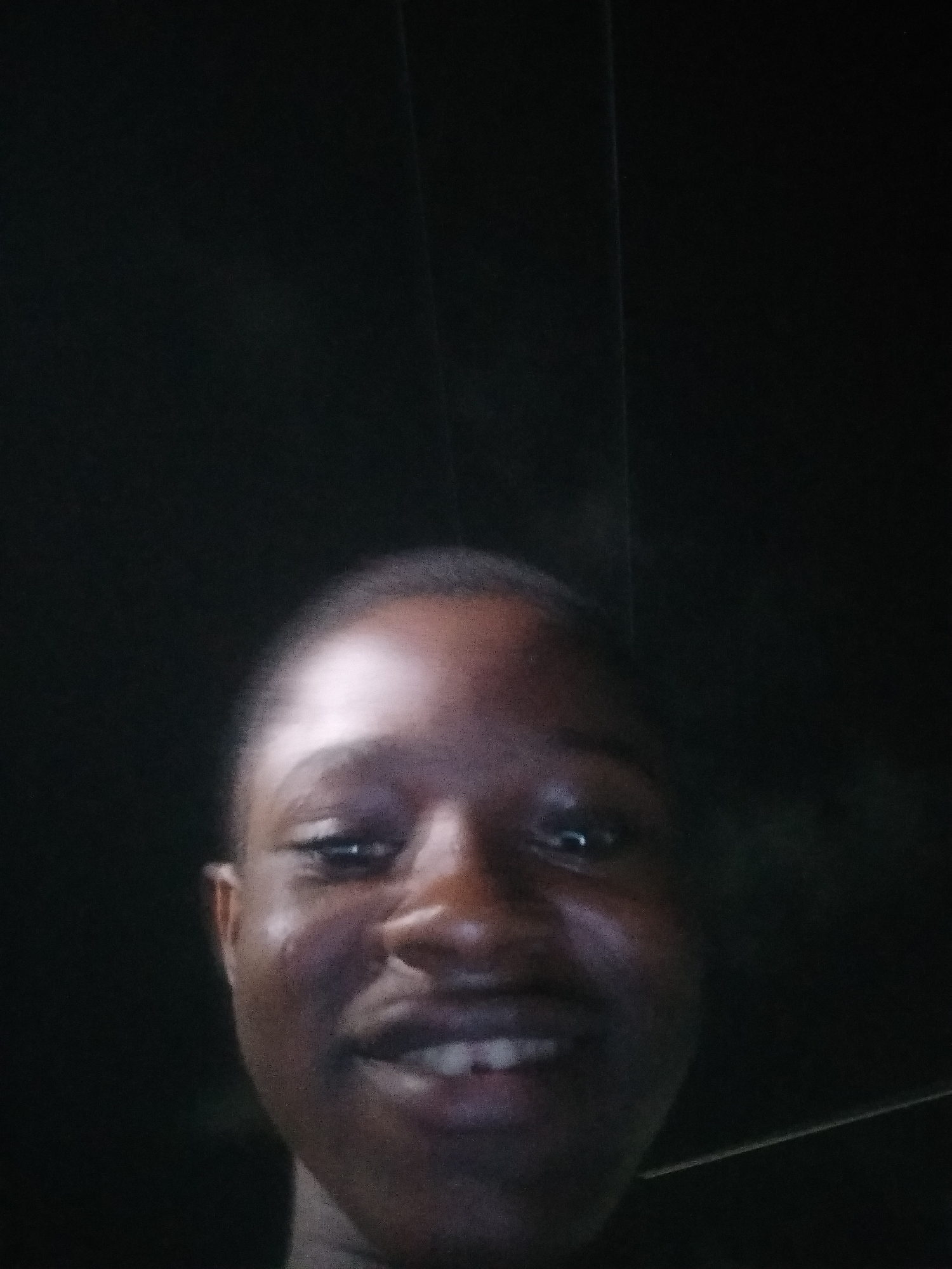 A close-up selfie captures a person with dark skin and short hair, smiling slightly. The background is uniformly dark and undefined, suggesting an indoor setting. The person's face is the primary focus, with details like eyes, nose, and mouth visible. No specific location or other people are visible.