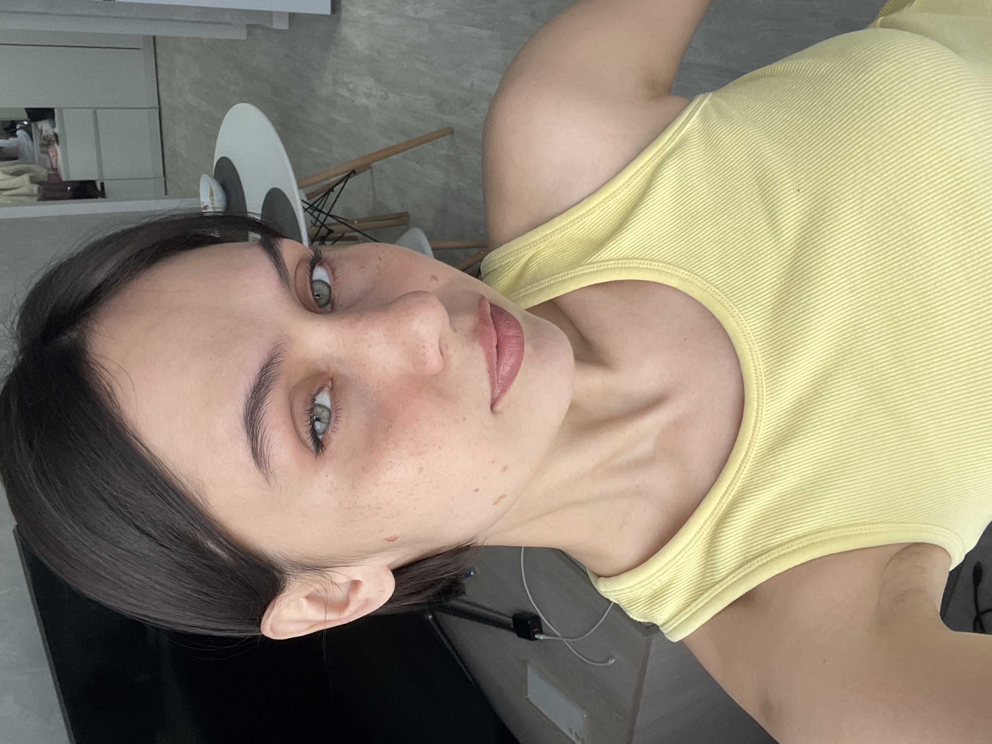 A woman with fair skin, dark brown hair, and light green eyes is looking at the camera, turned slightly to the side. She has freckles on her face. She is wearing a yellow ribbed tank top. In the background, there is a white chair, a white cabinet, and what appears to be a kitchen or dining area. It is not possible to determine the exact location in Izhevsk, Russia from this image alone.
