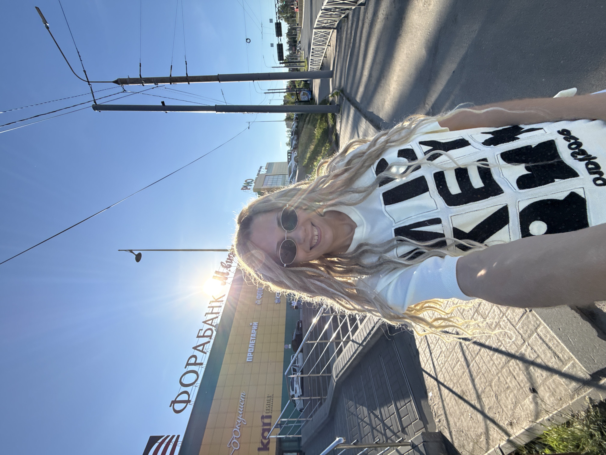 A female individual is captured in a selfie outdoors in Rusanovo, Russia. She has long, light blonde dreadlock-style hair, wears dark oval sunglasses, and is smiling directly at the camera. She is dressed in a light-colored, long-sleeved top featuring large black, block-like patterns that spell out "MICKEY" and include the text "DJD/2026" near the bottom right. Her right arm is extended to hold the camera.
The background on the left features a large commercial building with a yellow facade. Prominent Cyrillic signage on the building includes "ФОРАБАНК" (FORABANK), "ПРОЛЕТАРИЙ" (PROLETARIY), "ФОТО" (PHOTO), "документи" (documents), and "kari". A concrete ramp with metal handrails leads up to an entrance of this building. Several parked vehicles are visible in front of the building. The sun is low in the sky, positioned behind the building, creating a strong sunburst effect and lens flare, indicating either early morning or late afternoon.
To the right of the subject, a multi-lane road is visible, bordered by a metal pedestrian fence and a grassy embankment. Multiple utility poles, some equipped with streetlights and carrying numerous overhead wires, line the street. Another commercial building with "РИО" (RIO) signage is discernible in the mid-distance on the right. The sky is clear blue, and long shadows are cast across the paved sidewalk, consistent with the low sun angle.