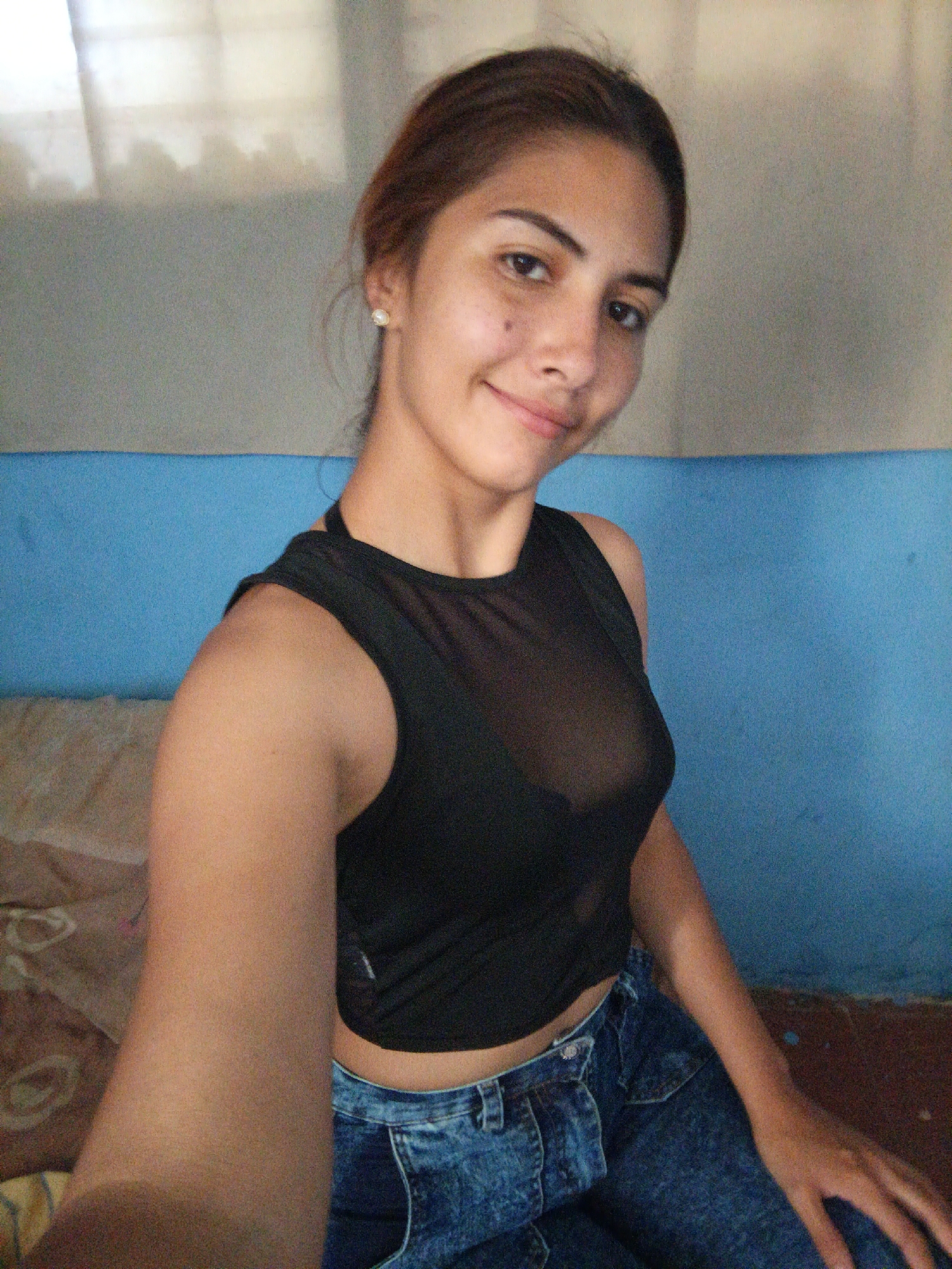 A young woman with auburn hair pulled back is captured in a selfie. She is wearing a black, sheer, sleeveless crop top and blue denim jeans. Her expression is a slight smile. In the background, a blue wall is visible, with a bed or couch on the left, covered with a patterned fabric. The lighting suggests it is daytime.
