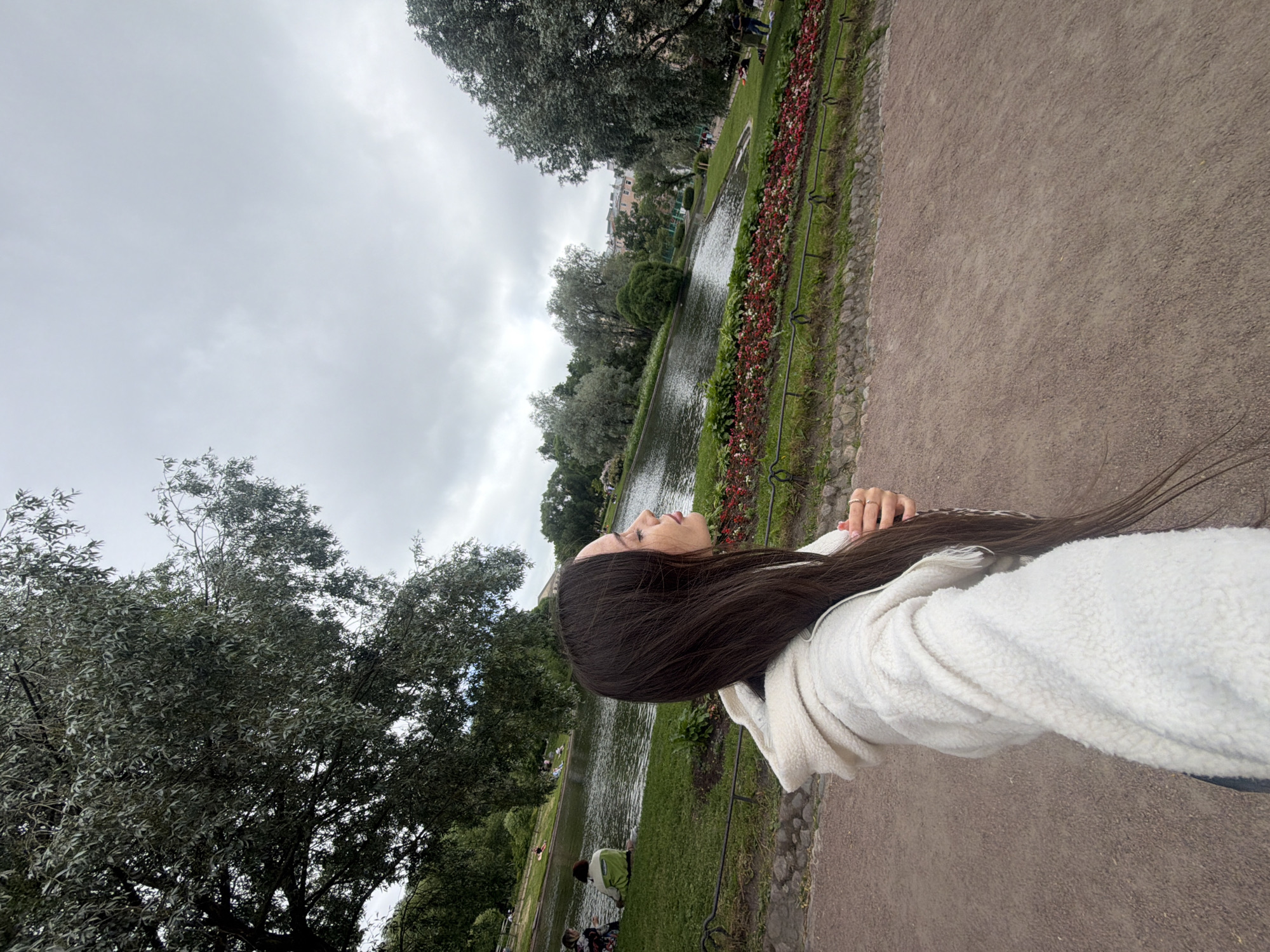 A woman is in the foreground, facing right, with her eyes closed and head tilted up. She has long, dark hair and is wearing a white sherpa-lined jacket. Behind her is a park with a pond, lush green grass, and trees. Several people are scattered throughout the park, some sitting by the water and others walking. The sky is overcast. The scene appears to be the Summer Garden in Saint Petersburg, Russia, a historic park established in 1704 by Peter the Great.