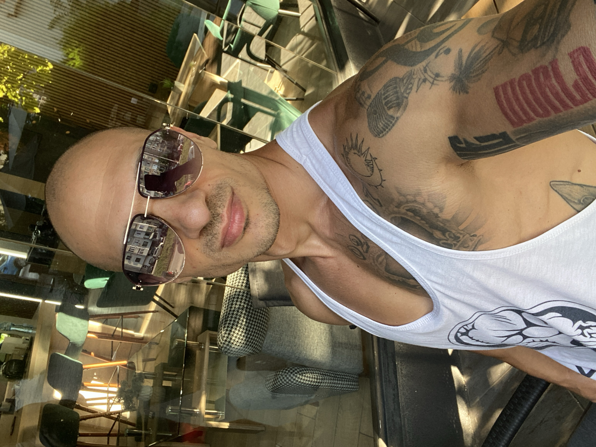 In Pleven, Bulgaria, a bald man wearing a white tank top and sunglasses takes a selfie in front of a glass-walled establishment. The reflection in the glass shows a row of high stools with green seats and wooden legs, a patterned armchair, and a glass table. The man has tattoos on his arms and chest, including a depiction of a microphone and a sunburst. The lighting suggests it is daytime, possibly late afternoon, with sunlight casting shadows and highlights. The overall impression is a casual, urban setting.
