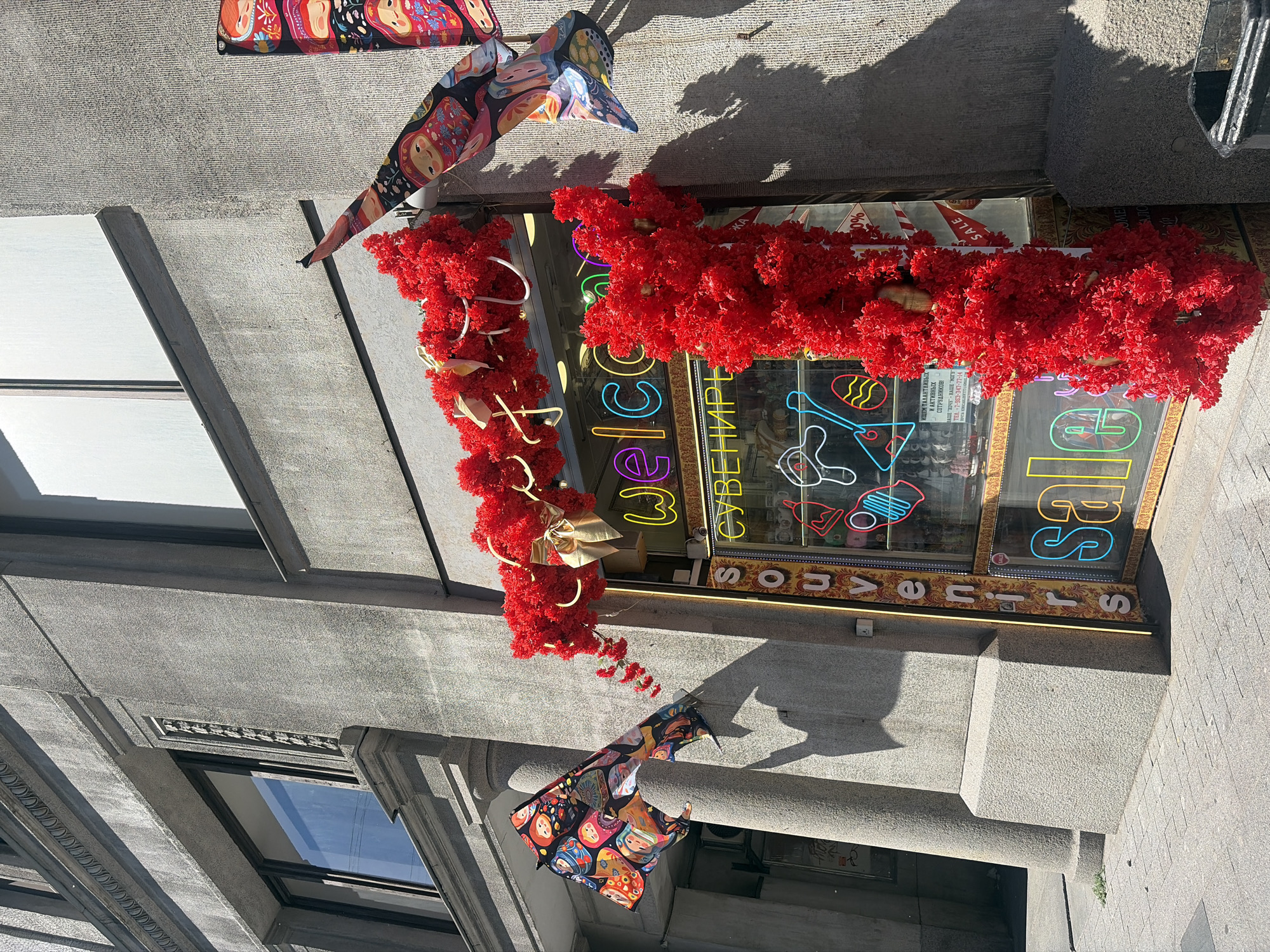 The image shows a store facade in Saint Petersburg, Russia, decorated with red floral garlands and a matryoshka doll motif. The store's window displays neon signs that read "welcome", "souvenirs", and "sale". The facade is made of grey stone, typical of the architecture found in the city. The sunlight casts strong shadows on the building and the sidewalk, indicating a bright day. There are no people visible in the image.