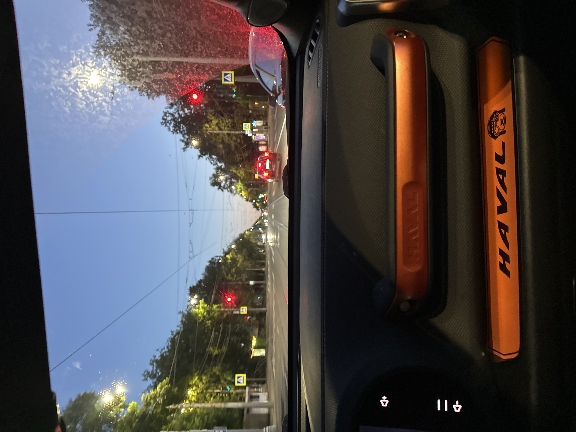 The image provides a view from inside a car, looking out the windshield at a street scene. The perspective is slightly tilted. The sky is a clear, light blue, suggesting it is either early morning or late afternoon/early evening. Trees line the street, some illuminated by streetlights, creating a warm glow. Traffic lights are visible, with some showing red, indicating traffic is stopped. Several yellow diamond-shaped signs with blue borders are also present, common traffic signs in Russia indicating warnings or directions. The road appears wet, hinting at recent rain or ongoing precipitation.
Inside the car, on the right side of the frame, two orange objects are prominently displayed. The one on the left is a handle or grip, with the faint imprint of "HAVAL" visible. The object on the right is longer and also orange, with the word "HAVAL" written vertically in black letters. A small circular emblem with a stylized animal head, possibly a lion or tiger, is also visible on this object. Below these objects, a dark dashboard display shows two illuminated icons, resembling a seedling or plant, and a musical pause symbol with two vertical bars.
The overall scene suggests a moment of pause in traffic, possibly at a red light. The combination of streetlights, traffic signals, and the interior of the car points to a metropolitan or suburban setting. The presence of the "HAVAL" branding on the objects suggests they are either accessories for a Haval vehicle or promotional items. The image captures a typical urban commute or travel scene in Nizhny Novgorod, Russia, during a time of subdued lighting conditions.