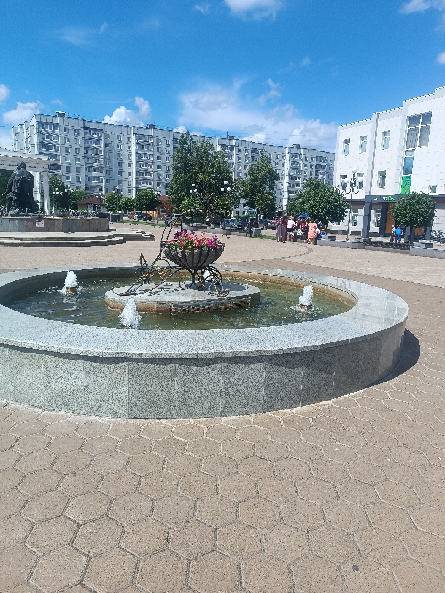 The image depicts a sunny day in a public square in Chaikovsky, Russia. In the foreground, a circular fountain with a granite rim features a decorative planter filled with pink and purple flowers. Water jets are active in the fountain. The ground surrounding the fountain is paved with hexagonal concrete tiles.
In the midground, a statue of a seated figure is partially visible on the left, suggesting a monument. Behind the fountain, several people are gathered, some standing and others sitting on a bench. A dark-colored statue on a pedestal is visible to the left of the fountain.
The background reveals a typical urban scene with multi-story residential buildings to the left and a modern white building with large windows and a green logo on the right. Trees line the square, providing some shade. The sky is blue with scattered white clouds. The overall impression is a clean and well-maintained public space.
