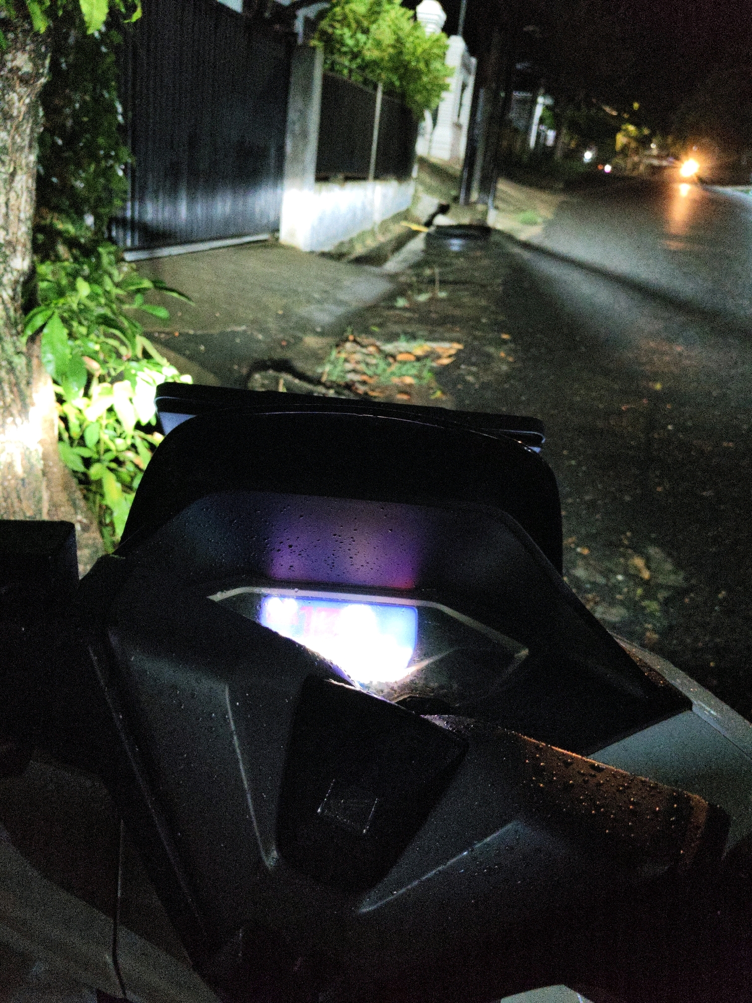 The image shows a close-up view of a motorcycle's digital display at night. The display is illuminated, showing a bright white and purple glow. Water droplets are visible on the black plastic surfaces surrounding the display, suggesting it may have been raining or the motorcycle is wet. In the background, a tree trunk with textured bark is visible on the left, with green foliage illuminated by a light source. Further in the background, a wet asphalt road can be seen, with distant streetlights and the headlights of an approaching vehicle. The environment appears to be a street in Lhokseumawe, Indonesia, given the context provided, likely near a residential or commercial area with a sidewalk and a fence. The scene is dark, with artificial lighting playing a significant role in illuminating the subject and its surroundings.