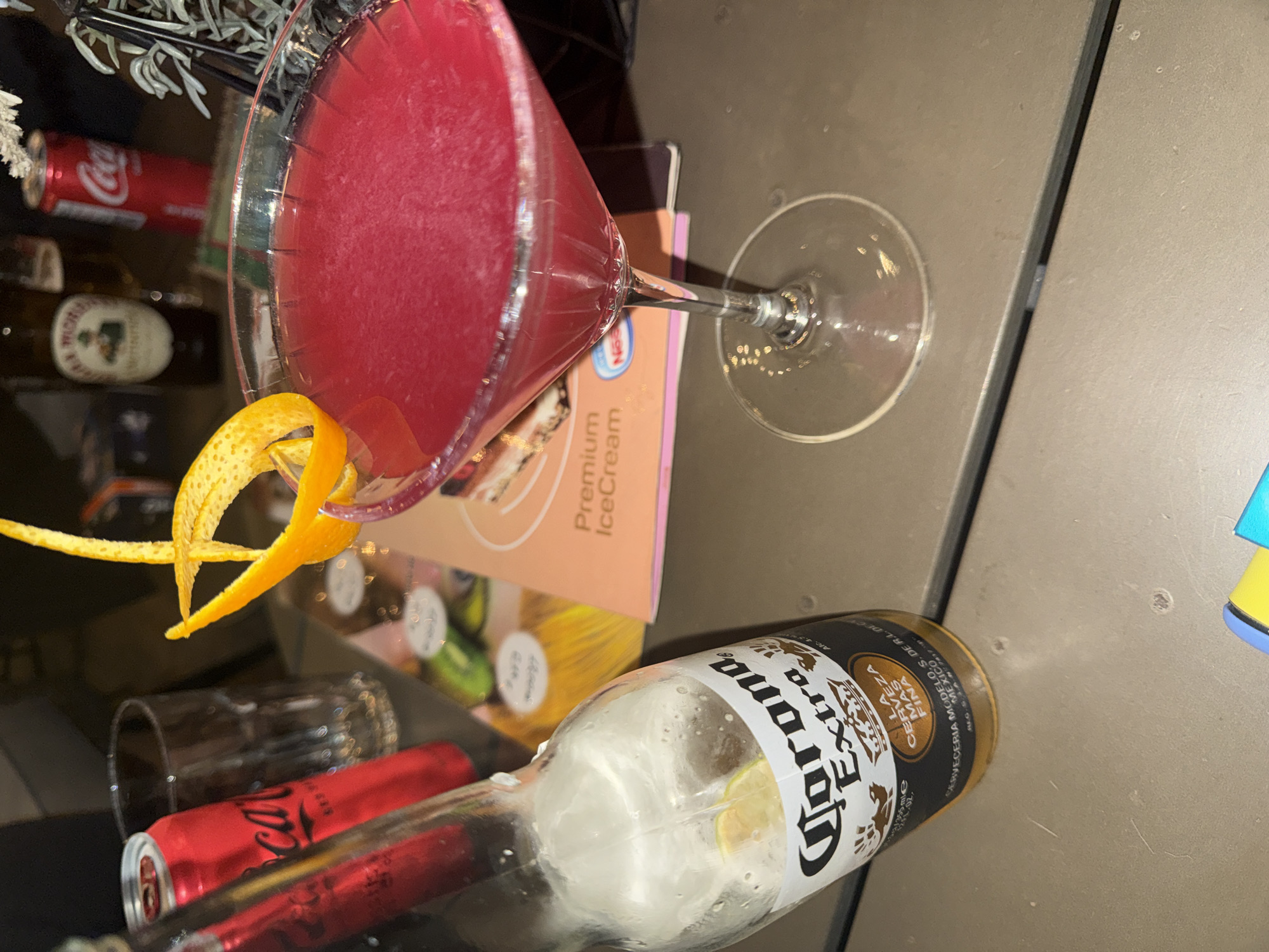 The image depicts an assortment of alcoholic and non-alcoholic beverages, with a vibrant pink cocktail in a martini glass as a central focus. The cocktail is garnished with a spiral of orange peel. Partially visible behind the glass is a container labelled "Premium IceCream".
Other objects present include a bottle of Corona Extra beer, a partially obscured Coca-Cola can, and what appears to be another beverage bottle in the background. The surface underneath is a neutral tone.
No people or actions are visible in the scene. The image lacks sufficient detail to determine its location in Pleven, Bulgaria.