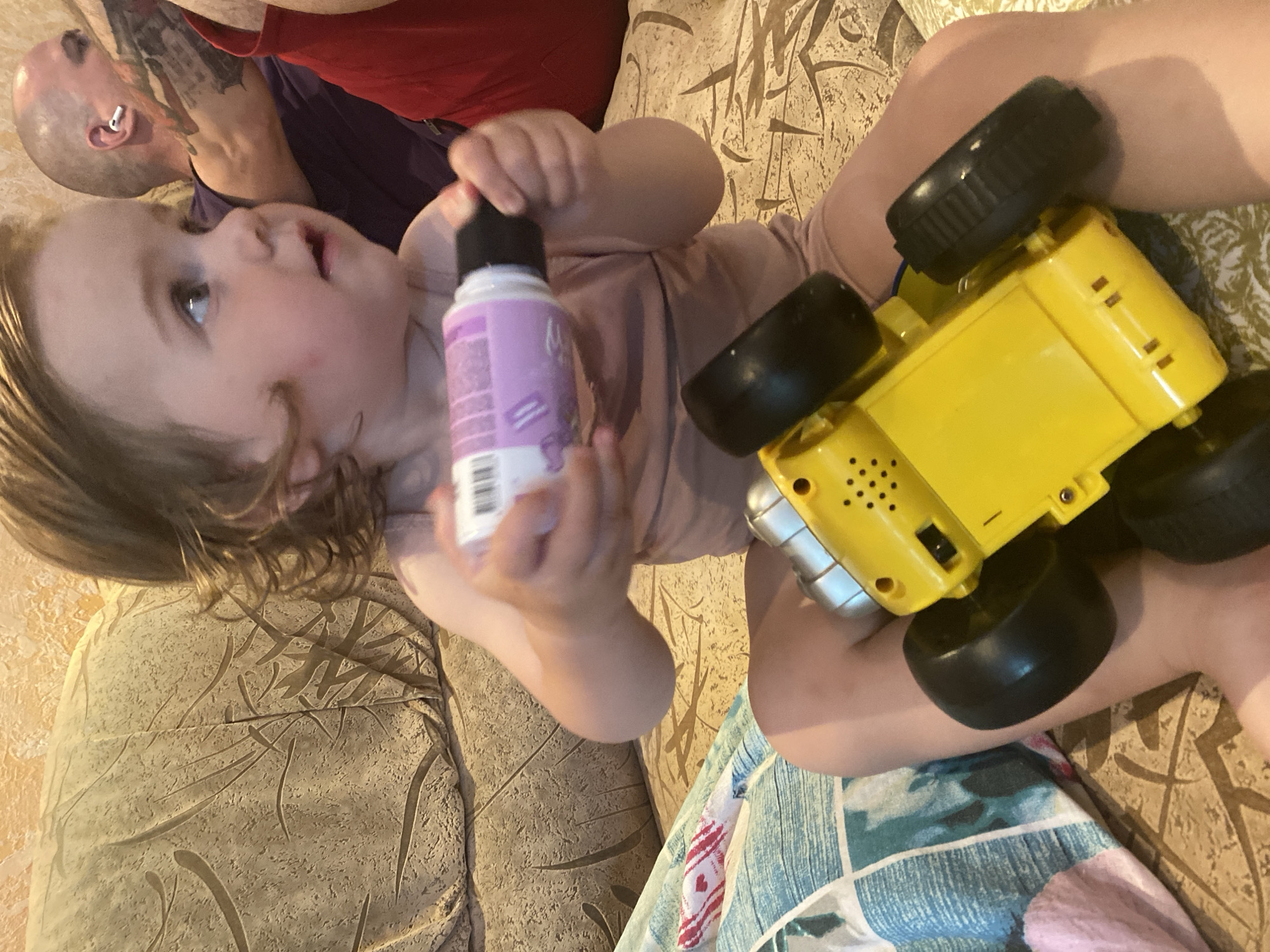 A young Caucasian child with light brown hair is sitting on a patterned sofa and holding a small, white bottle with purple writing and a black cap in their right hand. The child is wearing a light brown onesie. In their lap, the child is holding a bright yellow toy car with large black wheels. Behind the child, an adult Caucasian male with a shaved head and tattoos on his arm is visible, wearing a purple shirt and an earbud in his right ear. The sofa appears to be in a living room, with a textured wall in the background. There is a pillow with a blue and white patterned cover to the left of the child.