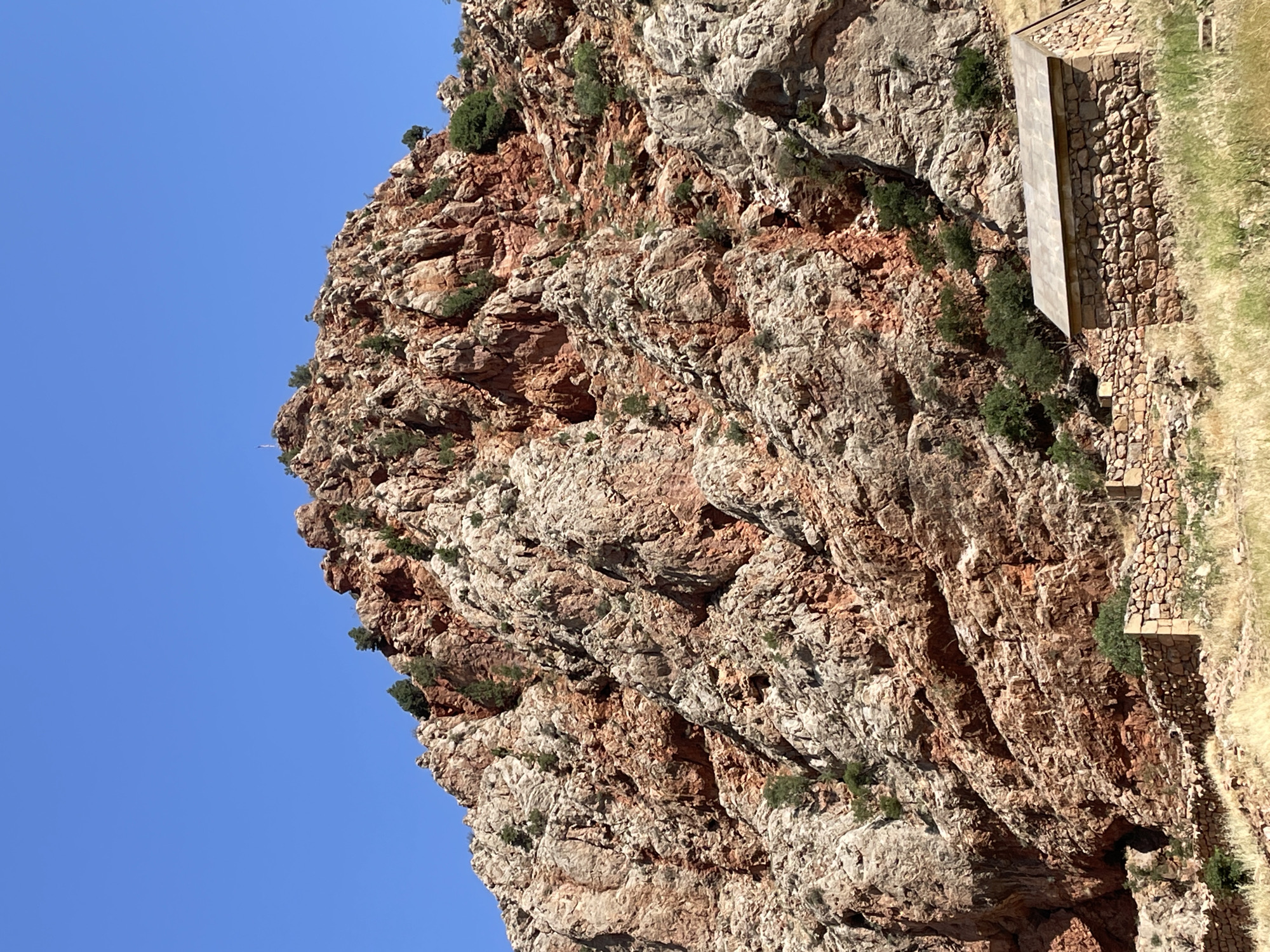 This is a photograph of a rocky mountainside in Areni, Armenia, under a clear blue sky. The mountain is composed of reddish-brown and gray rock formations, with sparse green vegetation growing in crevices. In the foreground, there are low stone walls and dry, yellowish grass. A small, stone building with a flat, tiled roof is situated in the lower right portion of the image. The building appears to be a historical structure. There are no people visible in the image.