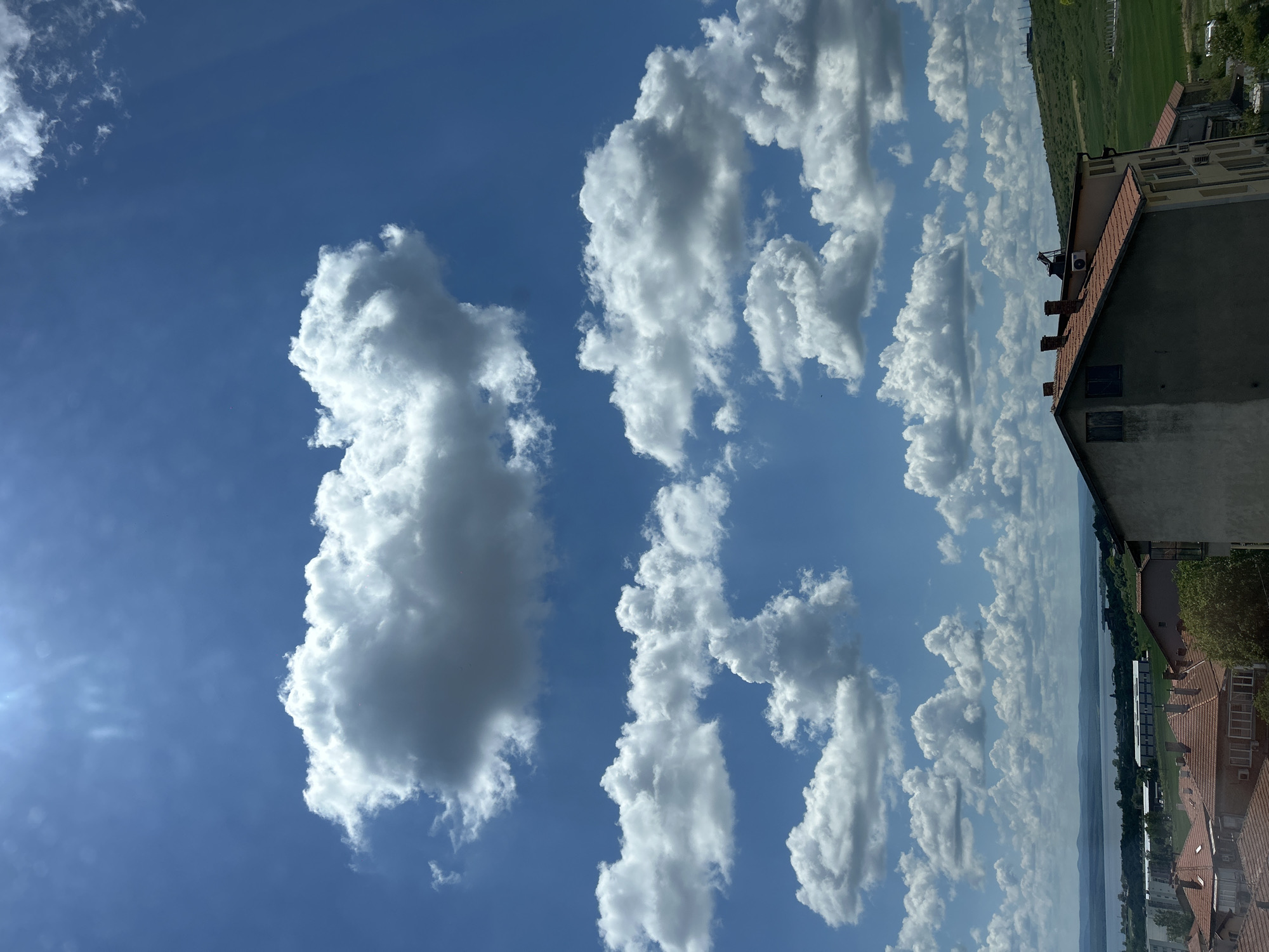 The image depicts a bright, partly cloudy sky above an urban area. The clouds are white and fluffy, scattered across a deep blue sky. The sun shines brightly in the lower left corner of the image. The lower right corner shows the edge of a building with a red tile roof, dark brick chimneys, and a light-colored side wall. Trees and greenery are visible in the background, along with rooftops of other buildings. A body of water stretches horizontally across the mid-ground. The photo appears to have been taken from an elevated position, possibly from a building's upper floor. The specific city location is not identifiable from this image.