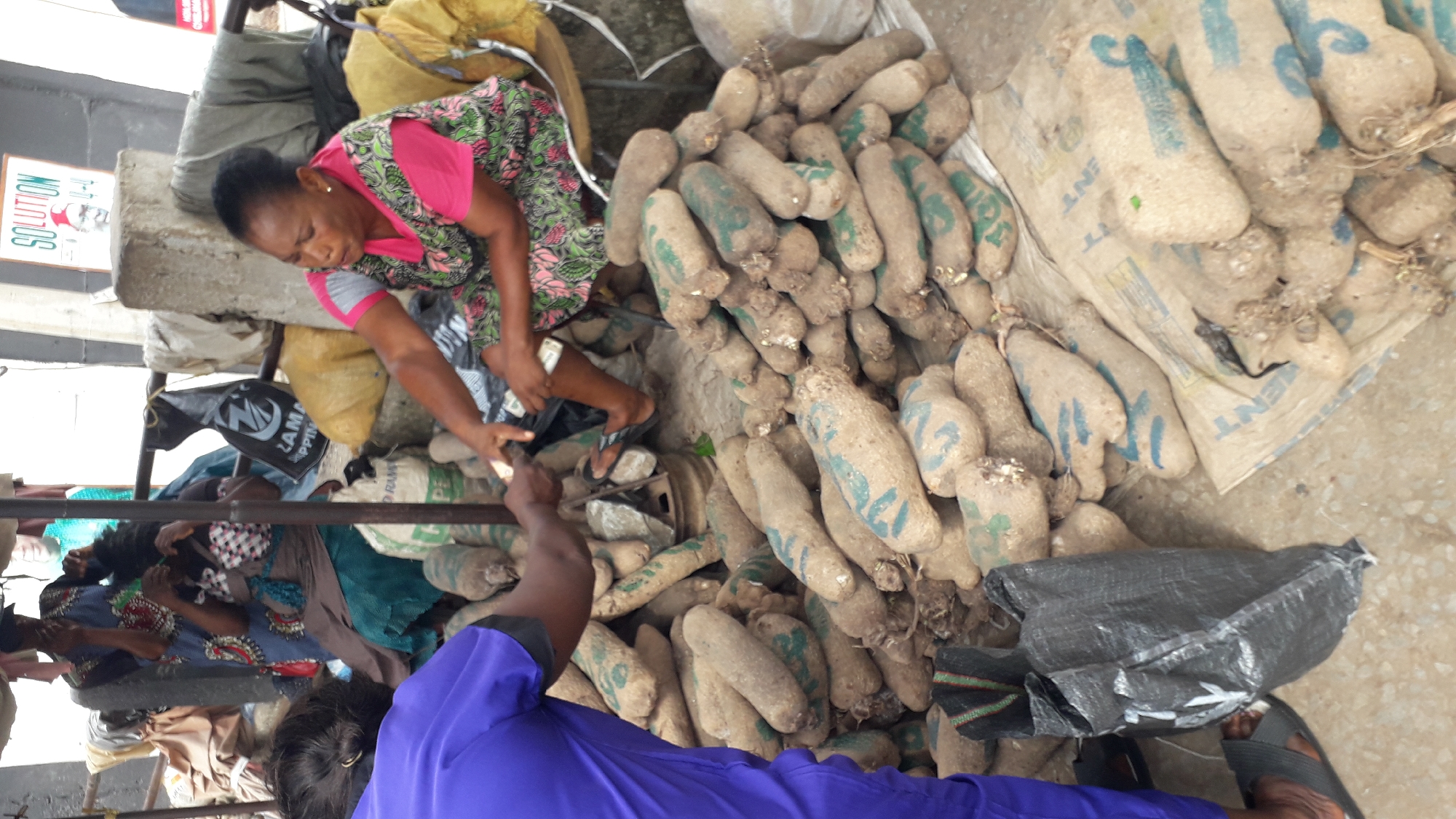 In a bustling outdoor market scene in Onitsha, Nigeria, a middle-aged woman in a pink floral dress and a vendor in a purple shirt are engaged in a transaction involving a large pile of yams. The yams, covered in dirt and marked with green or blue writing, are stacked on the ground and on a jute bag. The woman is reaching towards a black plastic bag, while the vendor appears to be handing over yams. In the background, other individuals are seated or standing, amidst bags and assorted items. The setting is a concrete surface, suggesting an established market area.