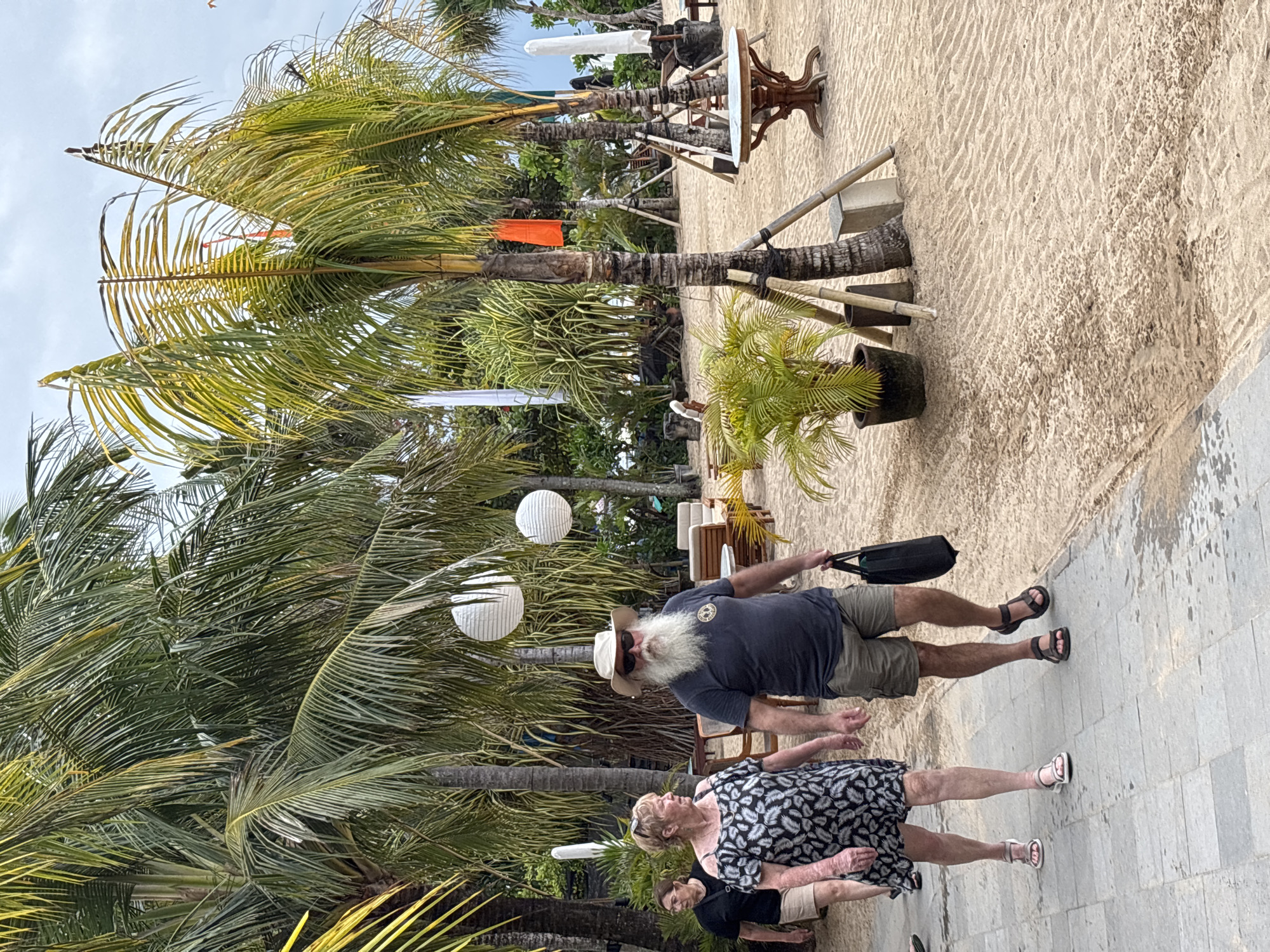 Two people, a man and a woman, walk on a paved path beside a sandy area lined with palm trees. The man, on the right, has a long white beard and wears a hat, sunglasses, a dark t-shirt, and cargo shorts. He carries a black bag in his right hand. The woman, on the left, wears a black and white patterned dress and sandals. A third person, a woman wearing a black t-shirt and shorts, is visible behind them on the path. The environment appears to be a tropical beach resort or walkway in Denpasar, Indonesia, with lush palm trees, sand, and outdoor furniture in the background. White spherical lanterns hang from a tree in the middle ground.