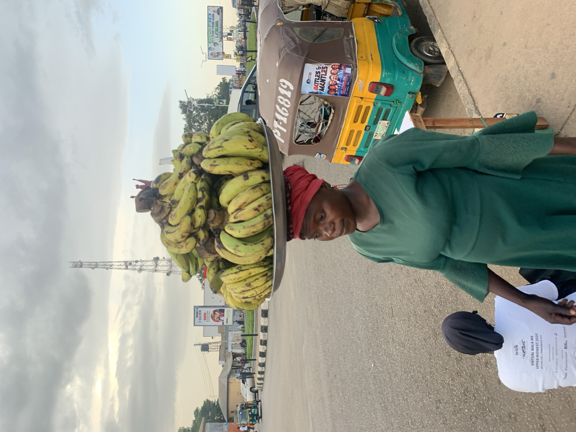 A woman with a headscarf and green dress balances a large tray of bananas on her head in Barakin Lerawi, Nigeria. The woman stands on a paved road, with a yellow and green auto-rickshaw parked to her right. The auto-rickshaw has a political advertisement on its side featuring a group of men and text that reads "Battles & Mantles" and "Let's Go Mutuwang Again LEMA". Behind the woman, a communication tower is visible against a cloudy sky. In the background, there are buildings, other vehicles, and pedestrians. Another person, wearing a white t-shirt with "WALK MS" printed on it, is partially visible in the lower left corner, facing away from the camera.