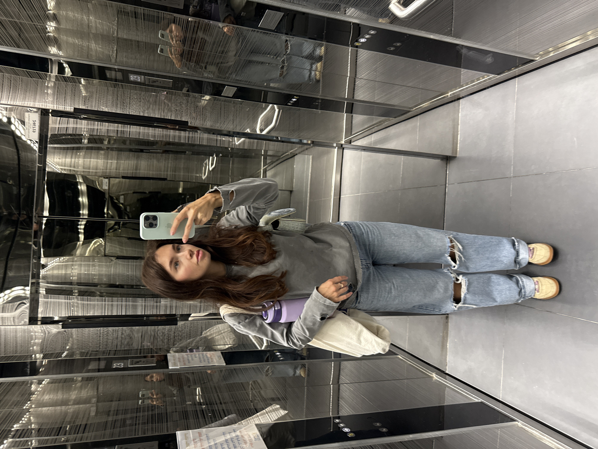 A young woman stands in a modern, mirrored elevator, possibly in Tel-Aviv, Israel. She is taking a selfie with a light green smartphone, holding it in her right hand. She has long, dark brown hair, a light complexion, and wears a grey sweatshirt, ripped light-wash blue jeans, and tan shoes. She is also carrying a purple water bottle and a white tote bag. The elevator features a grey tiled floor, stainless steel walls with reflective panels, and recessed lighting on the ceiling. In the reflections, multiple images of the woman can be seen, along with glimpses of elevator control panels and floor indicators. A sign with Arabic script is visible near the floor.