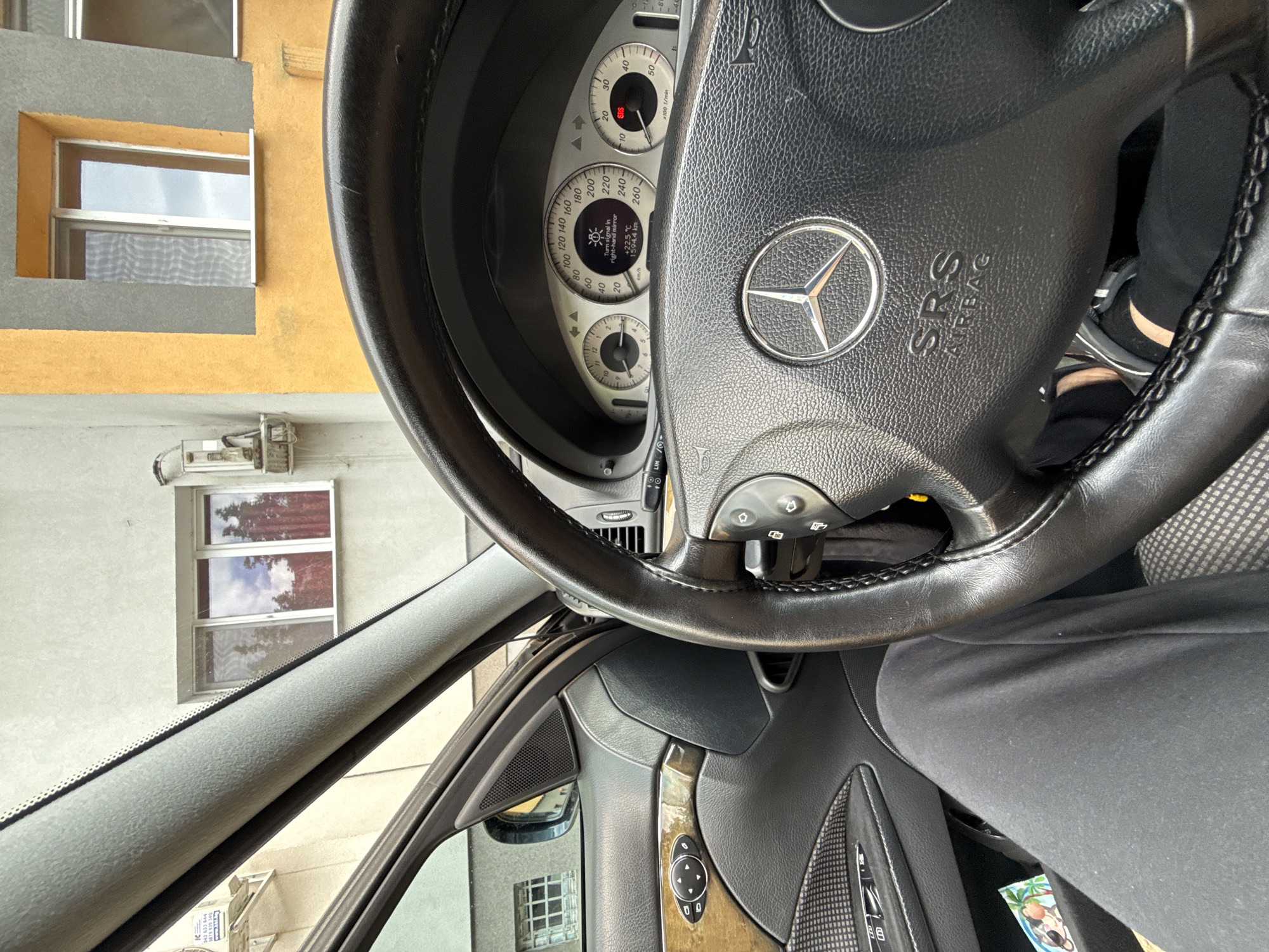 The image depicts the interior of a Mercedes-Benz car, specifically focusing on the steering wheel and dashboard. The steering wheel is black leather with a central Mercedes-Benz logo, labeled "SRS Airbag." The dashboard displays a speedometer and other gauges, showing a "Turn signal in right-hand mirror" message with "+22.5 °C" and "1594.4 km". Outside the car windows, buildings are visible. The OCR text includes numbers that might be related to contact information, suggesting the location is in Stara Zagora, Bulgaria. There is a person wearing black clothing in the driver seat.