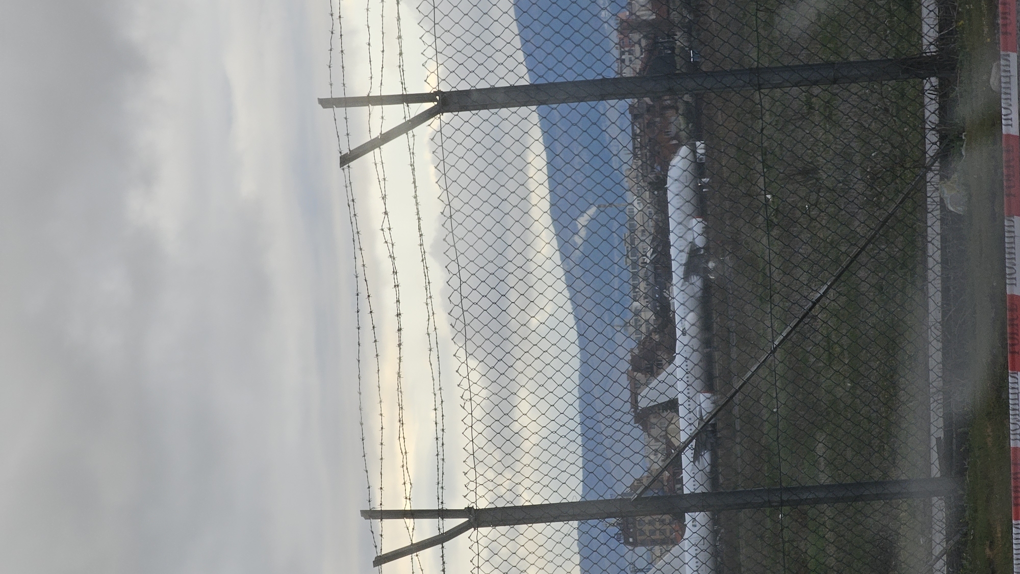 The image depicts a view through a chain-link fence topped with barbed wire. In the background, there's a cityscape partially obscured by the fence. A white pipe or structure is visible in the mid-ground on the right side of the fence. A red and white "ПОЛИЦИЯ" (Police) barrier tape is attached to the fence. The sky is overcast. Based on the "ПОЛИЦИЯ" tape, the location is likely in Bulgaria, with the city being Sofia due to the prompt's context. The scene suggests a restricted or secured area.