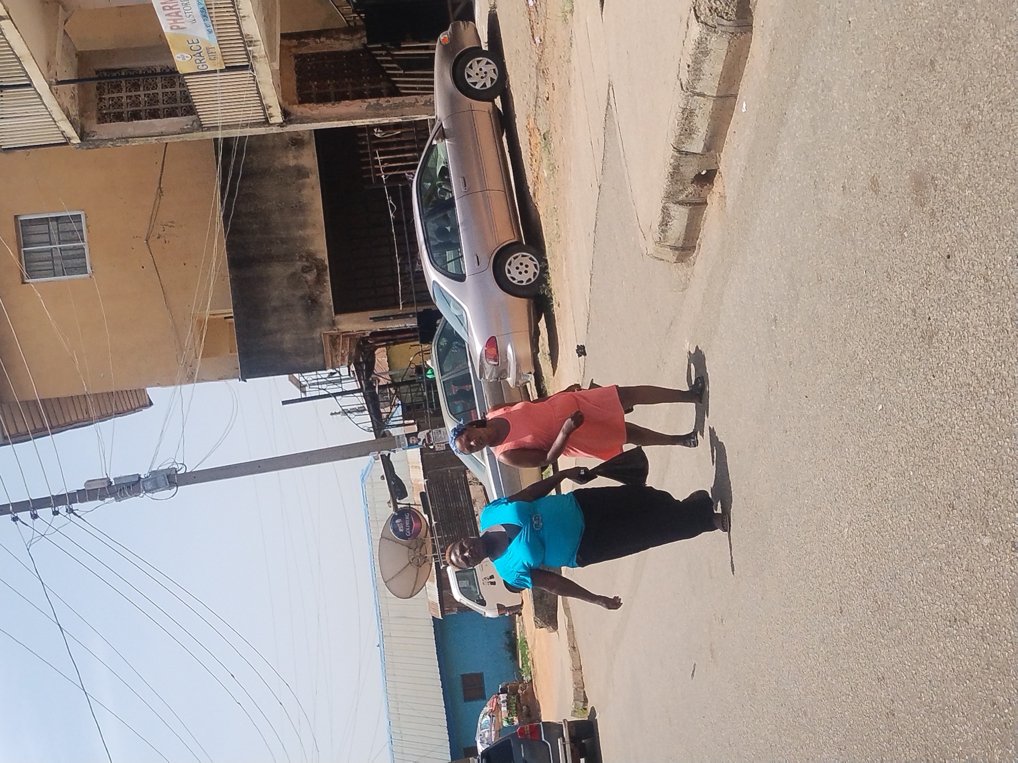 The image shows two women walking on a street in Jos, Nigeria. One woman is wearing a blue top and black pants, while the other is wearing a peach-colored dress. A silver car is parked on the side of the road. Power lines run overhead. Buildings are visible in the background, including one with a sign for "Grace Pharmacy."