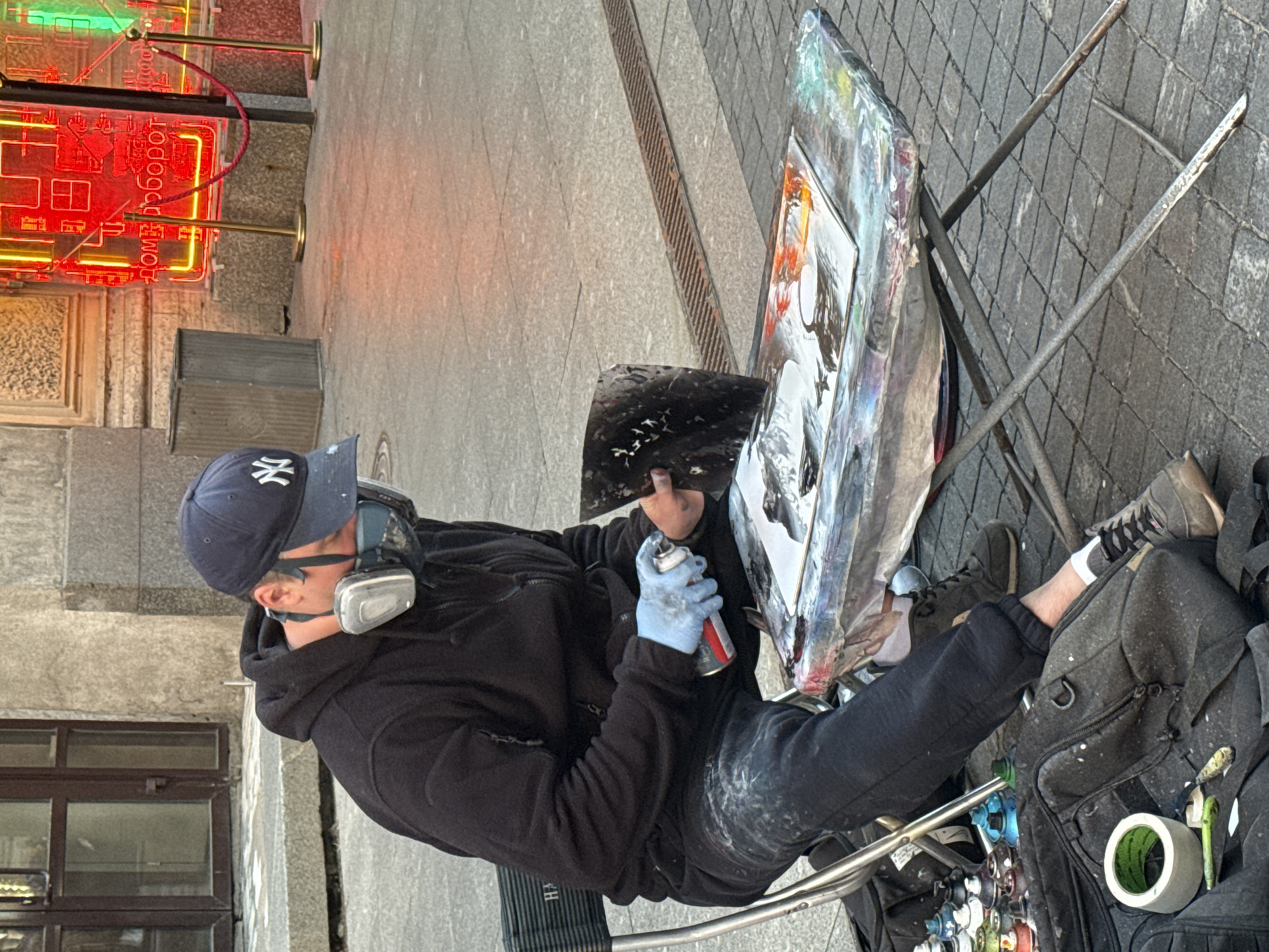 A male artist, wearing a black hoodie, black sweatpants, a blue baseball cap with a white New York Yankees logo, and a respirator mask, is seated on a folding chair. He is holding a spray can and a palette knife, actively working on a canvas on a folding table. The table is covered in paint splatters. In the background, a building with large windows and a neon sign with Cyrillic text is visible. The ground is paved with cobblestones. Numerous spray paint cans are arranged on the ground next to a black backpack, along with a roll of tape. The scene appears to be an outdoor urban environment, likely in Saint Petersburg, Russia, given the Cyrillic script on the neon sign.
