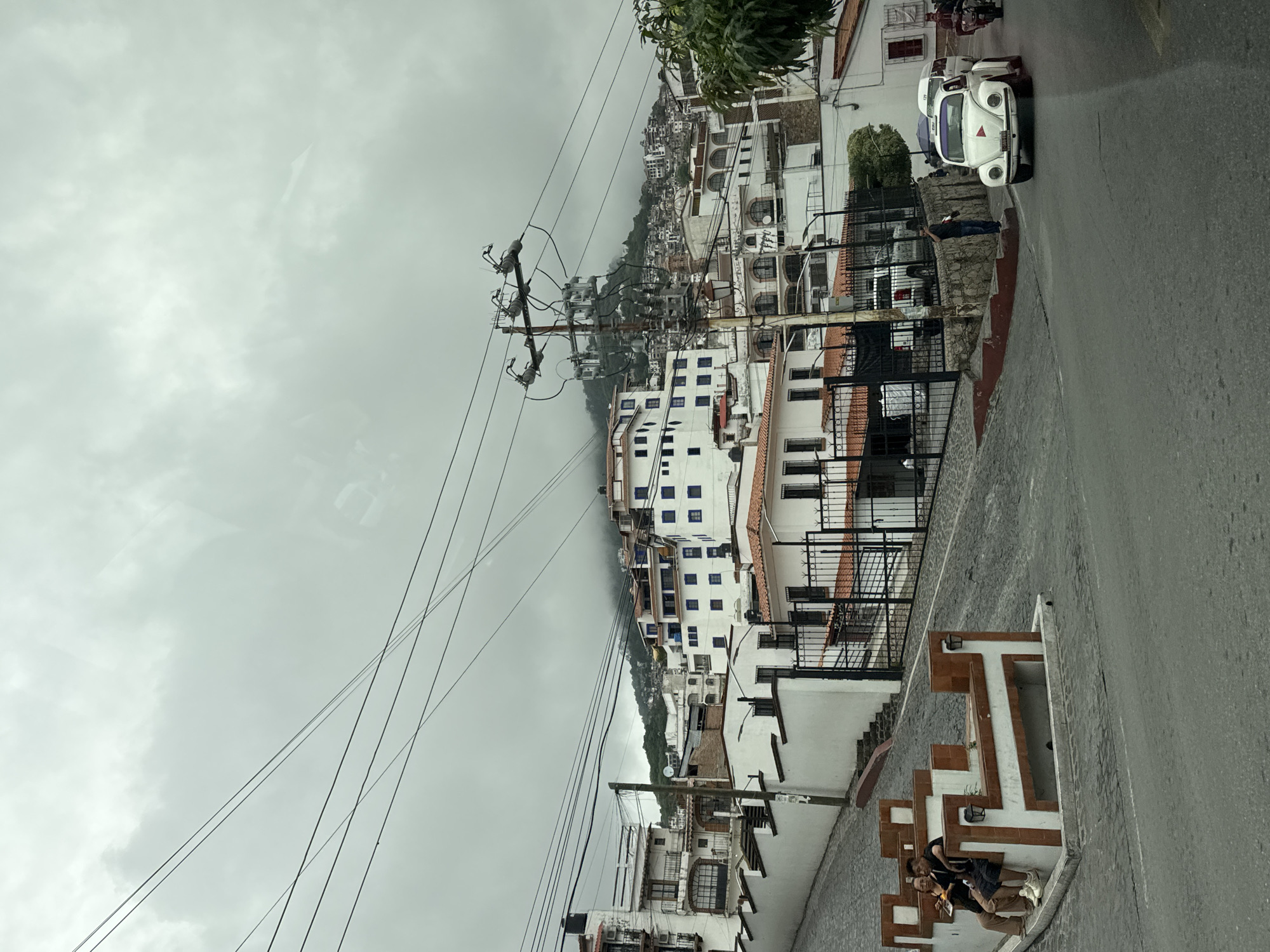 The image depicts a steep urban landscape in Taxco, Mexico, under an overcast sky with heavy grey clouds obscuring the upper reaches of the distant hills. Numerous black power lines crisscross the upper portion of the frame. Buildings are predominantly white with terracotta-colored tiled roofs, densely packed, and tiered along the inclining terrain. A prominent white building in the mid-ground features multiple small blue window frames.
In the lower-left quadrant, two individuals are seated on a decorative, multi-level public bench structure made of white and reddish-brown materials. One person wears a black shirt and dark shorts, while the other is in lighter-colored attire. They are observed looking down at an object in their hands.
A wide paved road occupies the foreground, transitioning to a cobblestone street that steeply ascends to the left. Along this cobblestone incline, a white wall with recessed sections runs parallel to the street. In the lower-right quadrant, a classic white Volkswagen Beetle, functioning as a taxi with "TAXI" and "175" marked on its roof light and a red triangular emblem on its hood, is positioned on the main road. Behind it, a white van is partially visible, and further to the right, a motorcycle with a rider is present. A lone individual in dark clothing stands next to a stone wall on the right side of the road, near the taxi.
A tall utility pole, equipped with multiple transformers and power lines, stands prominently in the mid-ground on the right, extending above the white buildings. Black metal gates are visible guarding entrances to properties on both sides of the main road. Patches of green vegetation are sparsely distributed, most notably on the far right and on the distant hillsides.