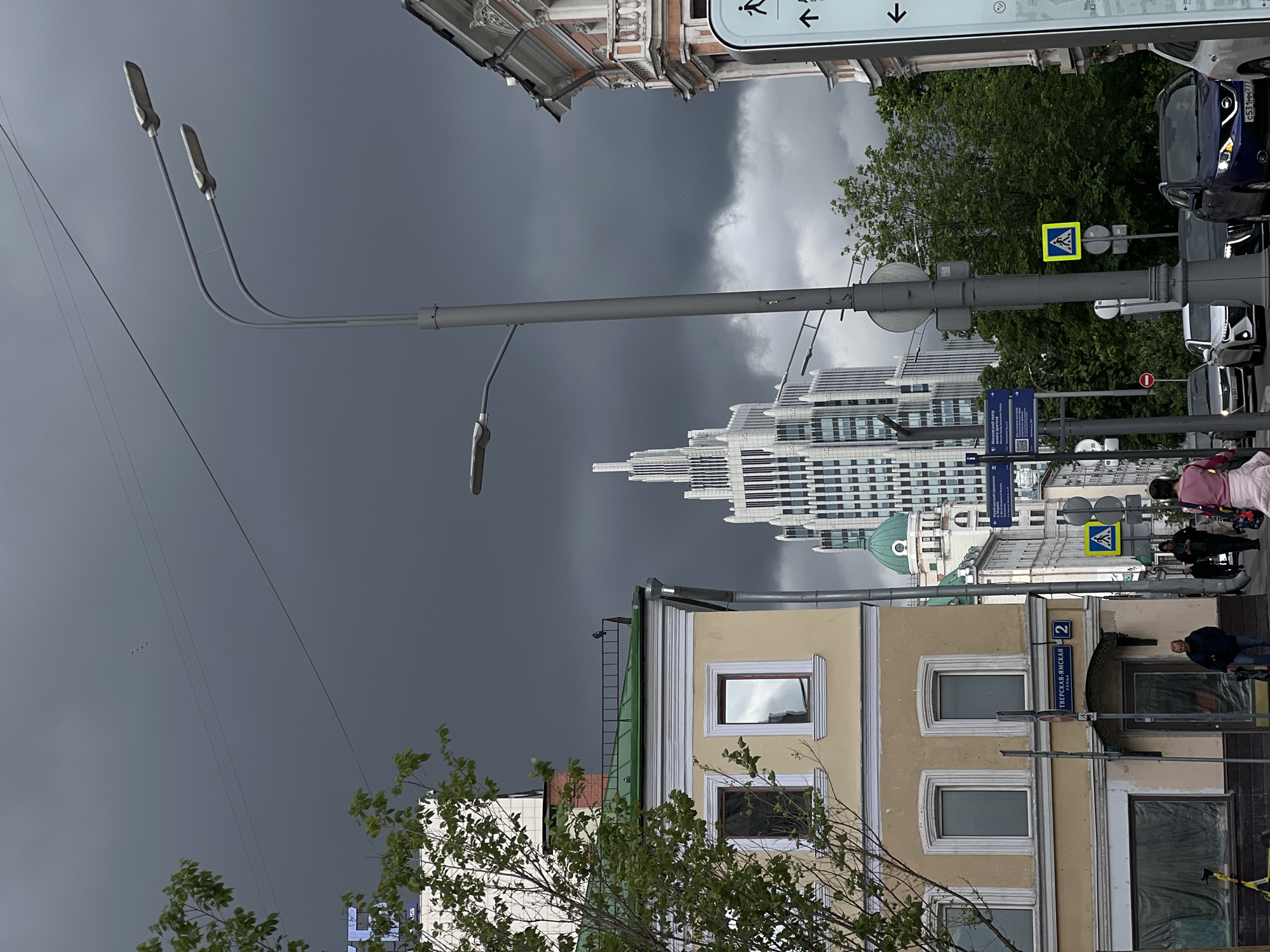 The image captures an urban scene in Moscow, Russia. A street view is framed by buildings, including a yellow multi-story structure with symmetrical windows and the Tverskaya-Yamskaya street sign, with the more modern white high-rise building of the White Square Office Center visible further back. Overhead, a dark, stormy sky looms. The street furniture includes a tall, gray lamppost with three curved lights, street signs indicating pedestrian crossings, and a bus route. Pedestrians are seen walking along the sidewalk, though the focus is not on them. Vehicles, including a dark-colored car, are parked on the right side of the street. The foreground features a tree with green leaves.