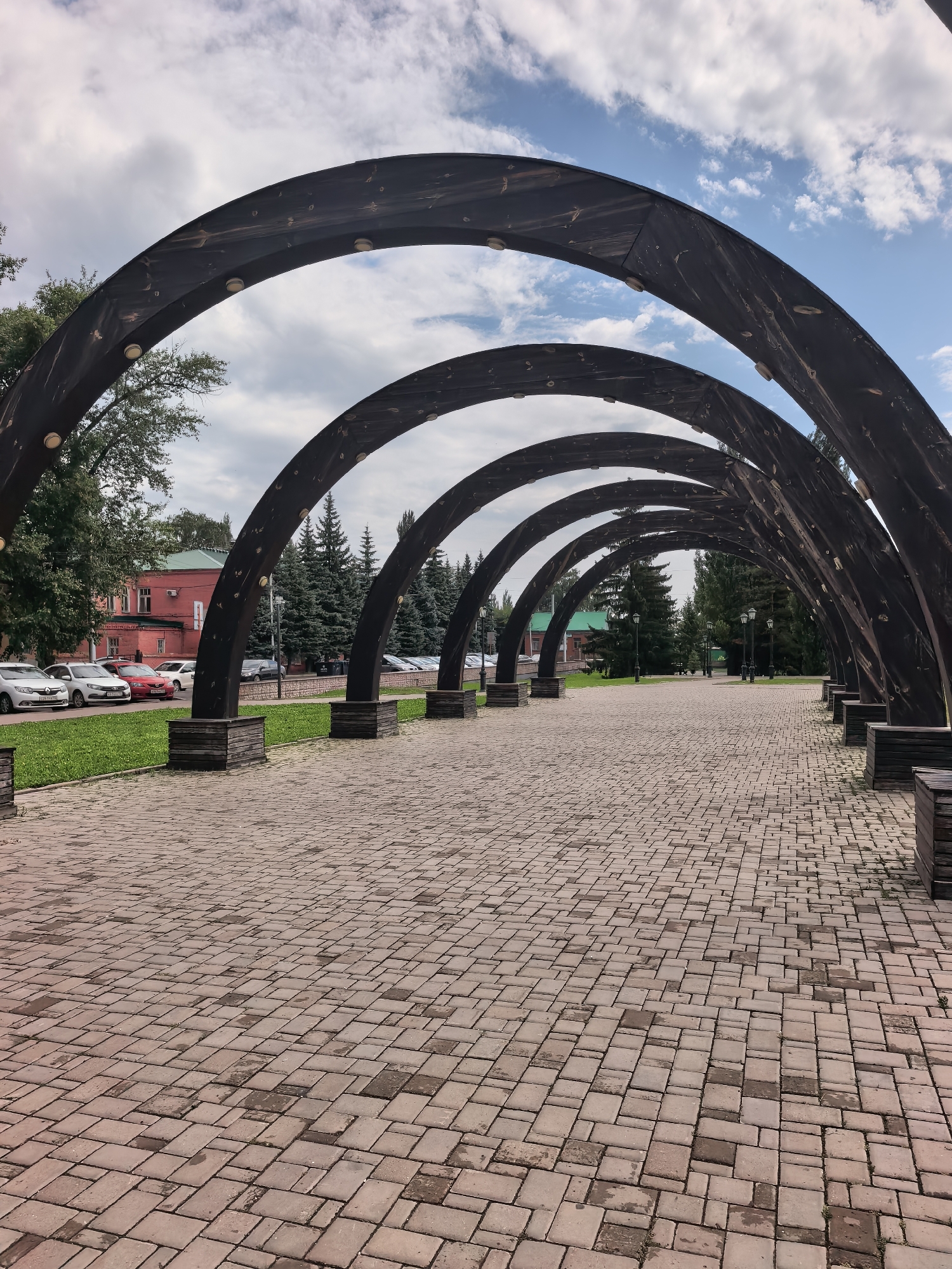 The image depicts a pathway lined with dark, arched wooden structures, creating a tunnel-like effect. The pathway itself is paved with reddish-brown rectangular bricks laid in a patterned fashion, with some areas appearing damp. On the left side of the pathway, there is a green lawn bordering a street with parked cars, including white sedans and a red car. Beyond the lawn and street, a red building with a green roof is visible, as are several tall, dark green evergreen trees. In the background, more evergreen trees and streetlights are scattered along the path. The sky is partly cloudy with patches of blue. This scene is located in Sterlitamak, Russia.