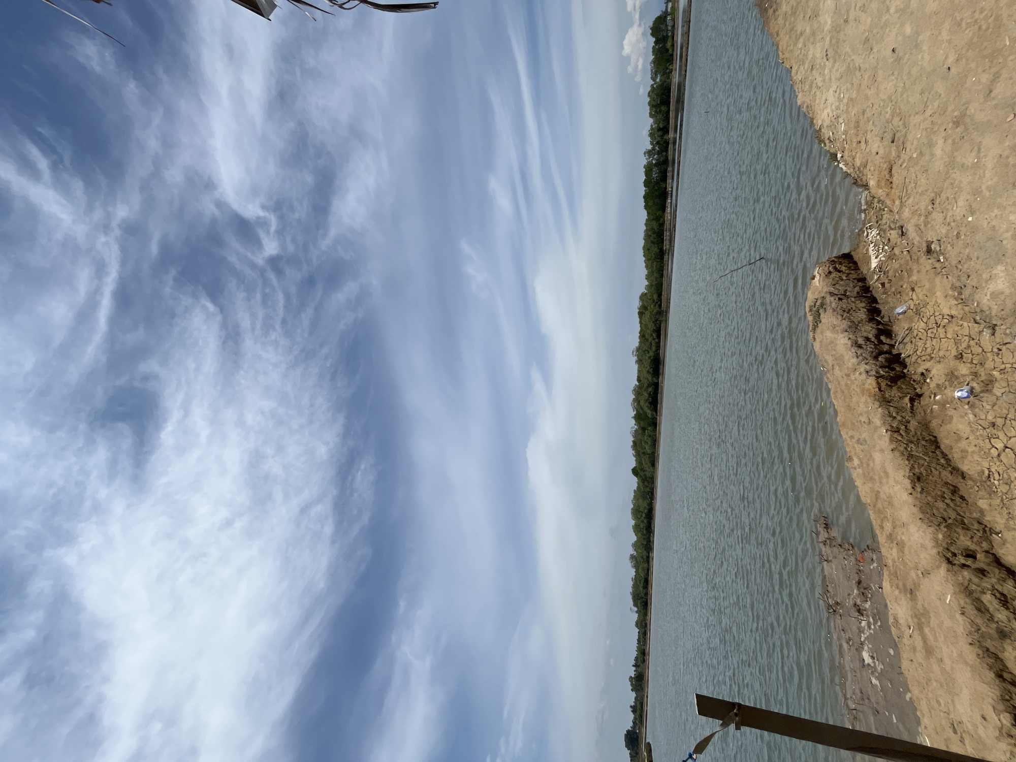 An outdoor, eye-level, full shot captures a tranquil body of water stretching towards a distant treeline under a bright, cloudy sky. The water is a muted green-grey, rippled by a gentle breeze. The foreground is dominated by a dry, cracked earth embankment on the right, transitioning to a muddy, debris-strewn edge closer to the water on the left. A metal pole with wires attached stands vertically on the left side of the frame, emerging from the murky shallows. In the distance, a dense, green treeline forms a horizontal band across the horizon, with hints of buildings or structures visible beyond. The sky above is a vibrant blue, crisscrossed with wispy, white clouds that appear streaky and elongated, suggesting wind. The overall scene evokes a sense of natural beauty and quietude, likely a coastal or estuarine environment. The image is located in Dayahtuha, Indonesia.