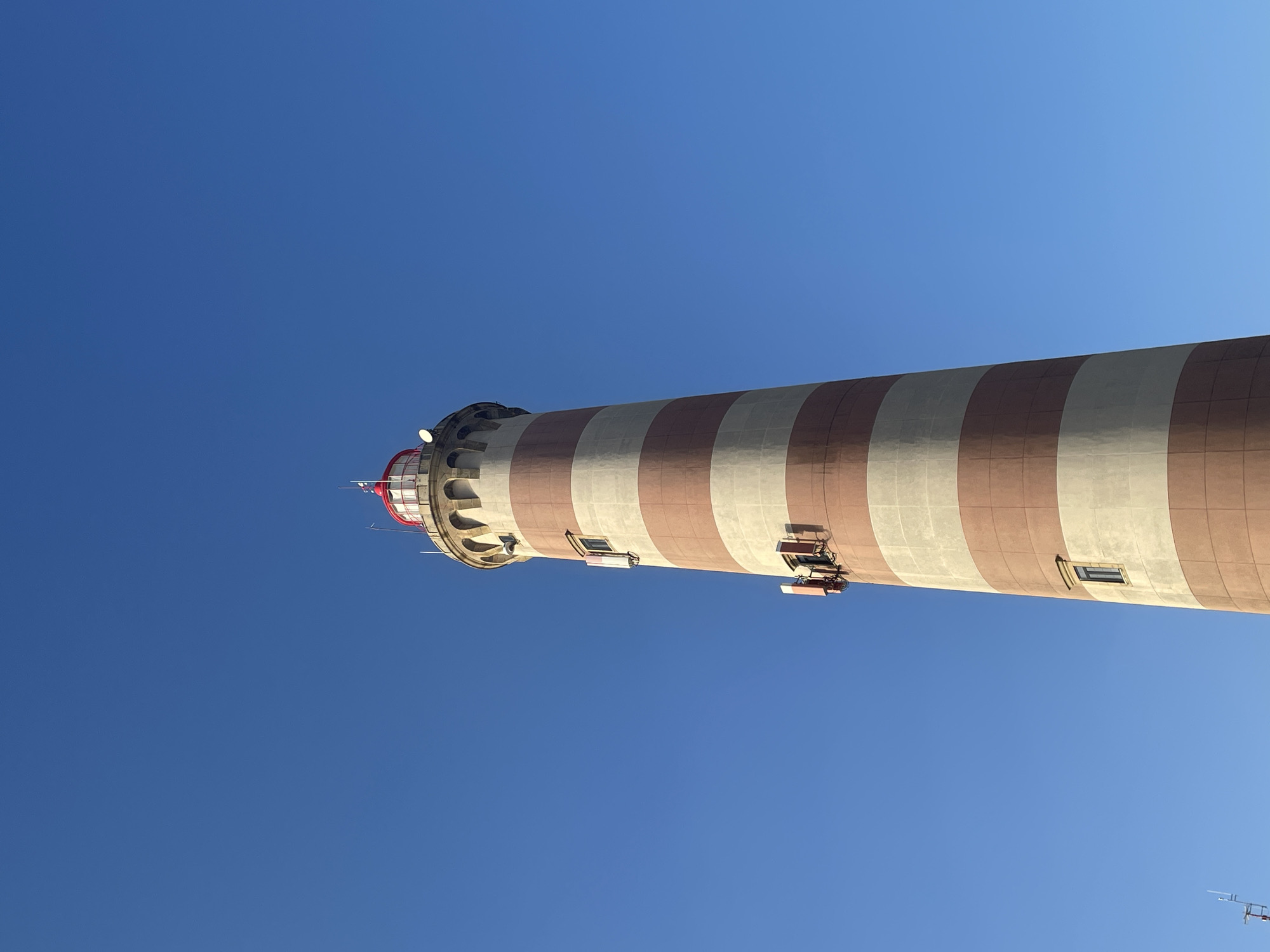 The image depicts the Barra Lighthouse in Gafanha da Nazaré, Portugal, under a clear, vibrant blue sky. The lighthouse is a tall, cylindrical structure with alternating horizontal bands of light beige and reddish-brown. It has a red-capped lantern room at the very top, surrounded by arched windows and a railing. Several small rectangular windows are visible along the body of the lighthouse, and on one side, there are multiple antenna-like structures attached to the exterior. The perspective is looking up at the lighthouse from a low angle, emphasizing its height against the vast expanse of the sky. There are no people or animals visible in the frame. The environment appears to be an open area, typical of a coastal location where a lighthouse would be situated.