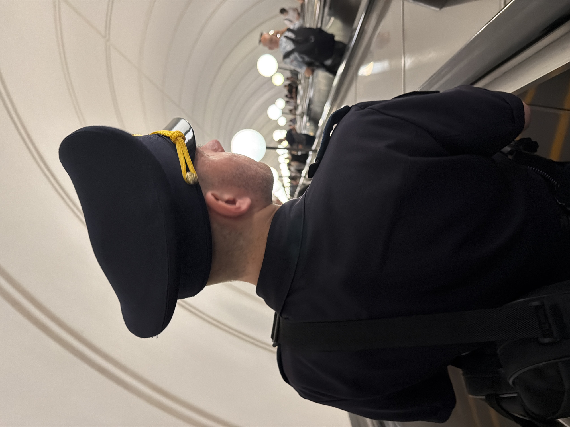 In this media file, the perspective is from behind and above a man wearing a dark uniform with a peaked cap adorned with a gold braid. He appears to be on an escalator in a Moscow metro station. The escalator is moving upwards, as indicated by the direction of other passengers visible in the background. The man's head is tilted back, and his gaze is directed upwards towards the arched ceiling of the station, which is illuminated by rows of bright, circular lights. The walls of the station are a light color, with subtle horizontal lines suggesting architectural detail. Other passengers are also visible on the escalator, some facing forward and others looking around. The overall impression is of a routine commute in a busy urban environment. The lighting suggests it could be daytime, but given it's a metro station, it's hard to determine the exact time of day. No text is visible in the image.
