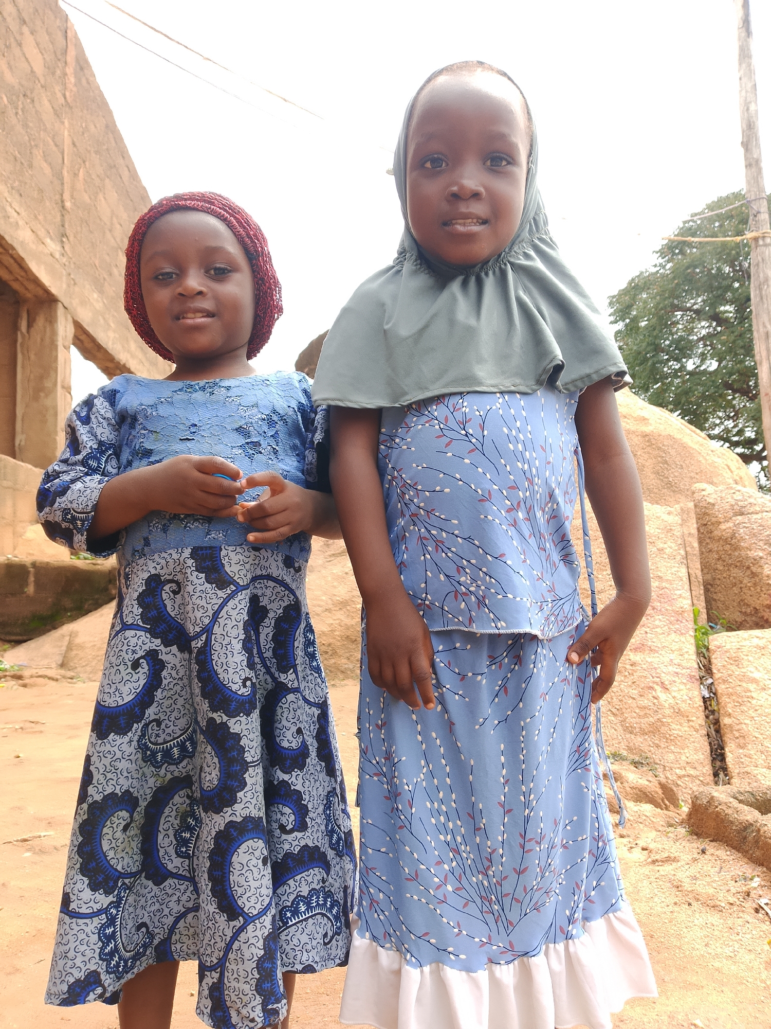 Two young girls are standing next to each other outdoors, with a weathered brick building to their left and a large pile of rocks to their right. The girl on the left, wearing a red head covering and a blue patterned dress, is holding a small object in her hands. The girl on the right is wearing a grey head covering and a blue dress with a floral pattern. Both girls are looking towards the camera and smiling. The ground appears to be dirt or sand. The sky is overcast.