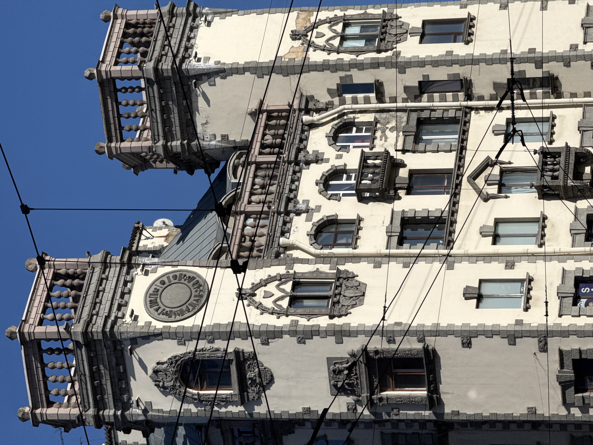 The image shows the facade of the Tolstoy House, located in Saint Petersburg, Russia. The architecture features elaborate window designs and decorative elements in varying shades of grey, complemented by the building's cream-colored walls. A series of balconies with ornamental railings are visible on the left. Power lines run across the view. The sky is a clear blue.