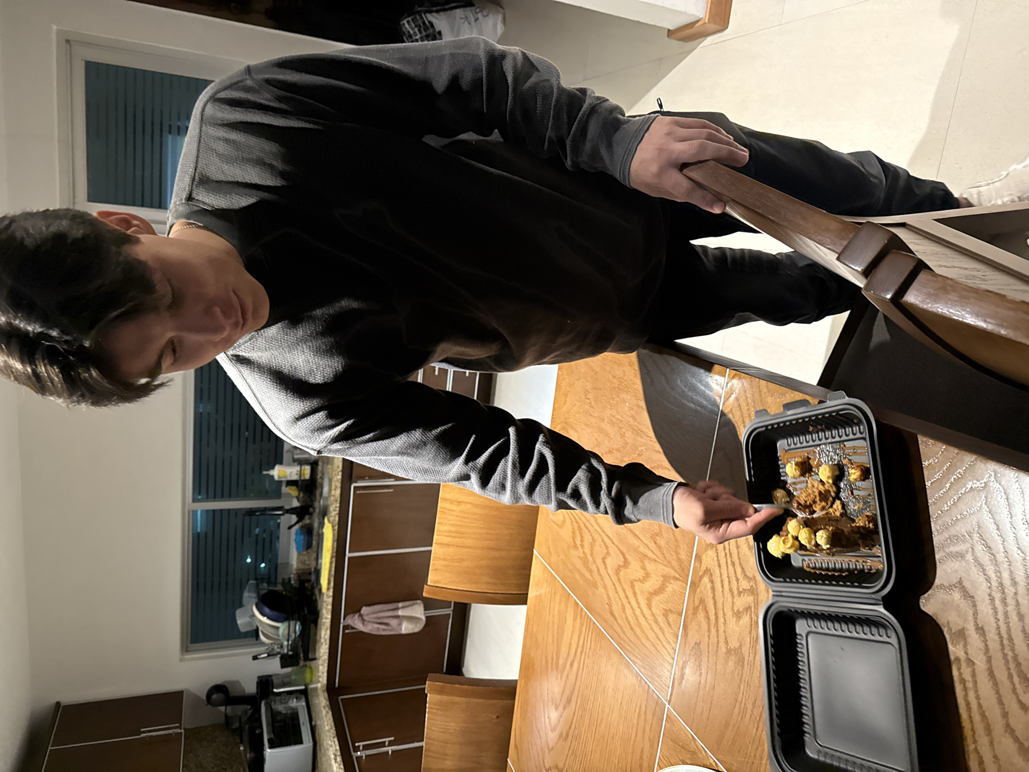 A person in a gray long-sleeved shirt is using a spoon to take food from a black take-out container. The container has two compartments, one with fried food and the other empty. The food consists of round yellow items mixed with brown sauce. The scene is indoors, on a light brown wooden table. Visible in the background are kitchen cabinets, a window with blinds, and a white wall. The location is San Bernardino Tlaxcalancingo, Mexico.