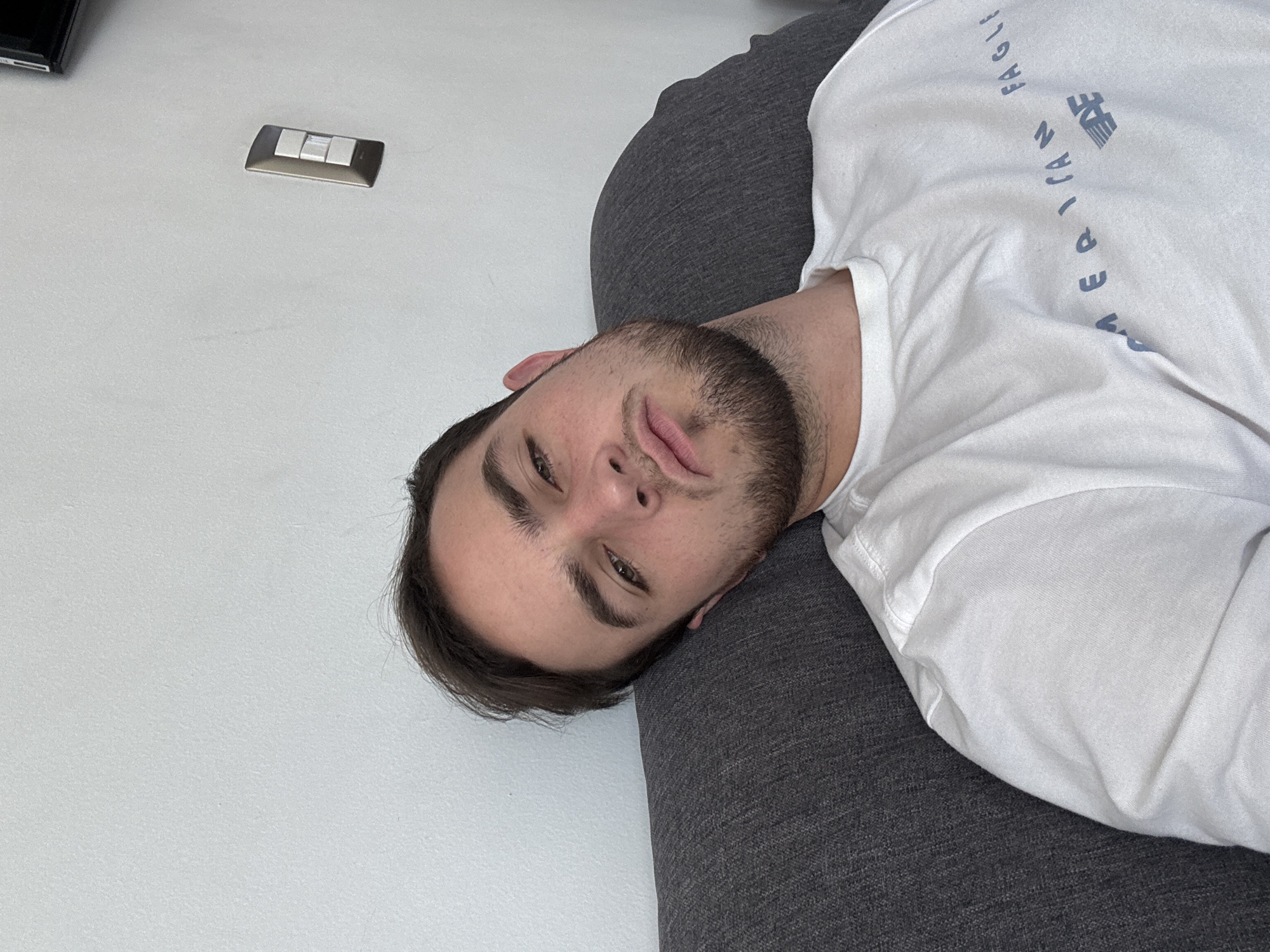 The image, rotated counter-clockwise, features a young man lying down indoors, resting his head on a dark gray upholstered surface, likely a sofa or cushion. He has dark hair, a neatly trimmed beard, and looks directly at the camera with a neutral, slightly pensive expression, his lips slightly parted. His eyes appear to be a shade of green or hazel.
He is wearing a white short-sleeved t-shirt. Text on the shirt, oriented vertically due to the image's rotation, reads "AMERICAN EAGLE," with portions of both words clearly visible.
The background consists of a plain, light-colored wall, which could be white or a very light gray. Mounted on this wall, in the upper left section of the original image, is a light switch with three rectangular white buttons encased in a metallic frame, possibly dark silver or bronze.
The lighting in the scene is bright and even, indicating indoor artificial light or diffused natural light, with no harsh shadows. The overall setting suggests a relaxed indoor environment, such as a living room or bedroom, located in San Bernardino Tlaxcalancingo, Mexico. The man's posture and direct gaze suggest he is either posing for the photo or captured in a moment of rest.